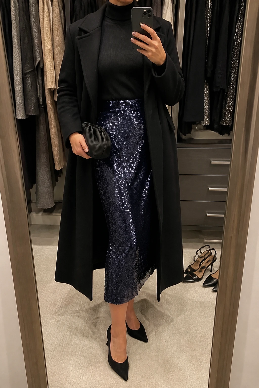 Image Prompt: Mirror selfie of a woman wearing a black fitted cashmere turtleneck sweater tucked into a midnight navy sequin midi skirt, black suede pointed-toe kitten heels, a long black tailored wool coat worn open, a small black satin clutch, and delicate silver stud earrings, holding a phone in front of a mirror, cropped at the NECK so the face does not show, with a relaxed casual selfie pose, in a walk-in closet mirror, warm moody indoor lighting, an open closet with clothes visible and a pair of heels on the floor nearby, realistic navy sequin reflections without exaggerated sparkle, natural cashmere texture and elegant party styling, latest iPhone photo quality, no additional clothing or accessories.