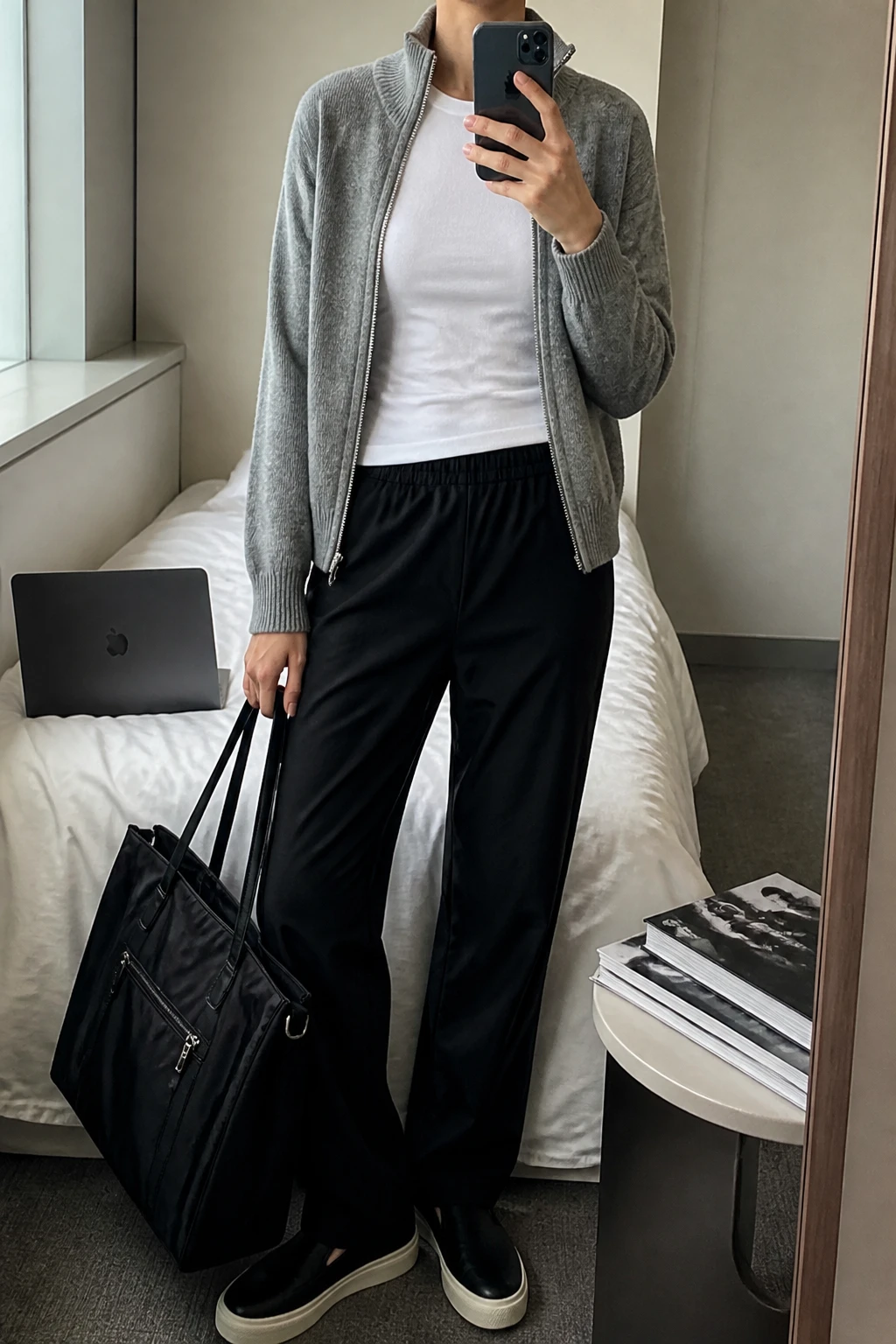 Image Prompt: Mirror selfie of a woman wearing a heather grey fine-knit zip-up cardigan with a high collar over a fitted white cotton crewneck T-shirt, black relaxed straight-leg stretch travel trousers with an elastic waistband, black cushioned leather slip-on sneakers, and a large structured black nylon zippered tote bag, holding a phone in front of a mirror, cropped at the NECK so the face does not show, with a relaxed casual selfie pose, in a minimal dressing room, soft natural window light, a laptop on the bed and a stack of fashion magazines on a side table, realistic grey knit texture, natural black trouser folds and understated airport styling, latest iPhone photo quality, no additional clothing or accessories.