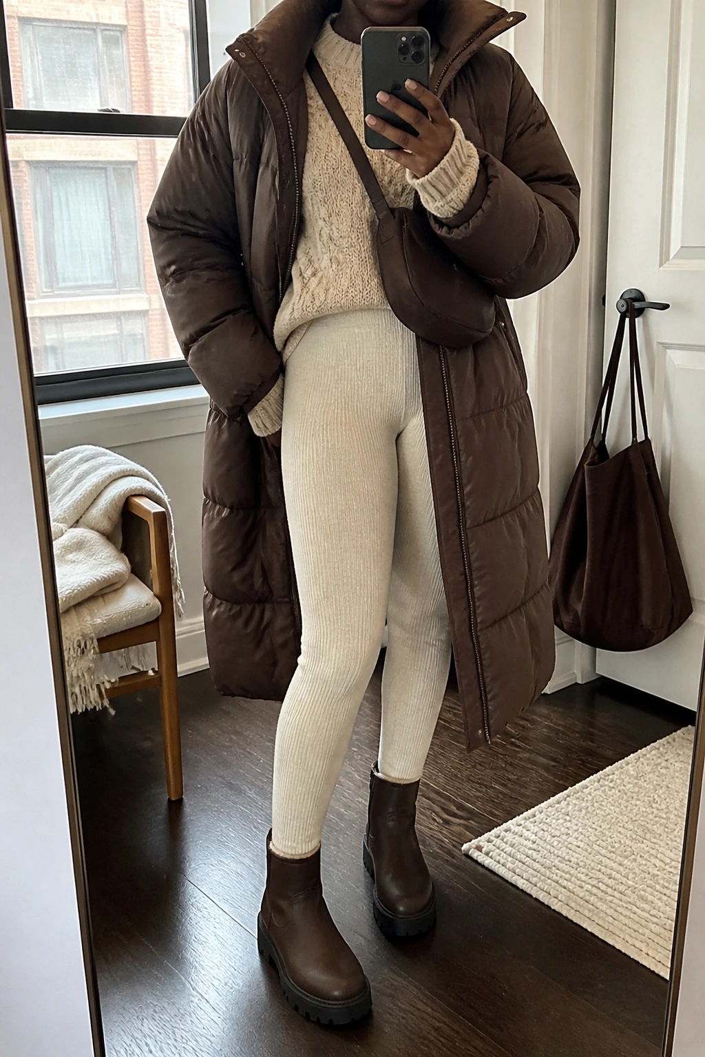 Image Prompt: Mirror selfie of a black woman wearing a long chocolate brown insulated quilted puffer coat over an oatmeal oversized thick cable-knit sweater, cream high-waisted fleece-lined ribbed knit leggings, dark brown waterproof leather chunky ankle boots with rubber lug soles, and a small dark brown nylon crossbody bag, holding a phone in front of a mirror, cropped at the NECK so the face does not show, with a relaxed casual selfie pose, in a stylish city apartment, soft natural window light, a folded blanket on a chair and a handbag hanging on a door handle, realistic chocolate puffer volume, natural cream knit fabric and cozy winter layering, latest iPhone photo quality, no additional clothing or accessories.