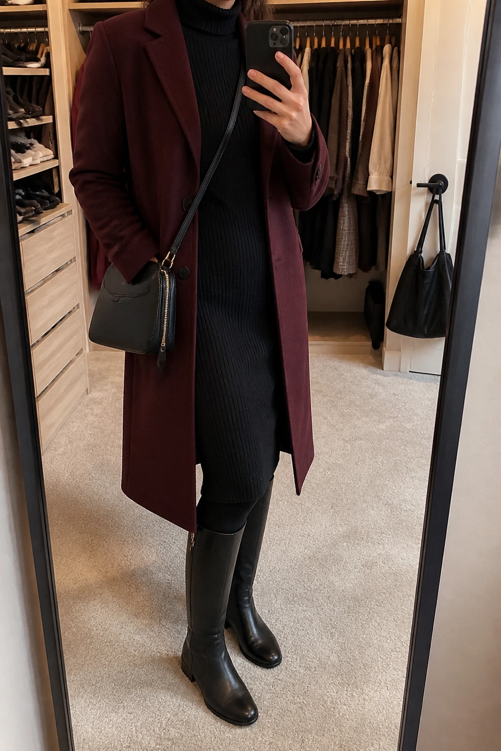 Image Prompt: Mirror selfie of a woman wearing a long burgundy tailored wool coat over a black long-sleeve ribbed knit turtleneck midi dress, black opaque fleece-lined thermal tights, black waterproof leather knee-high boots with flat rubber soles, and a small black leather zippered crossbody bag, holding a phone in front of a mirror, cropped at the NECK so the face does not show, with a relaxed casual selfie pose, in a walk-in closet mirror, warm soft indoor lighting, an open closet with clothes visible and a handbag hanging on a door handle, realistic burgundy wool texture, natural black knit dress folds and practical winter styling, latest iPhone photo quality, no additional clothing or accessories.