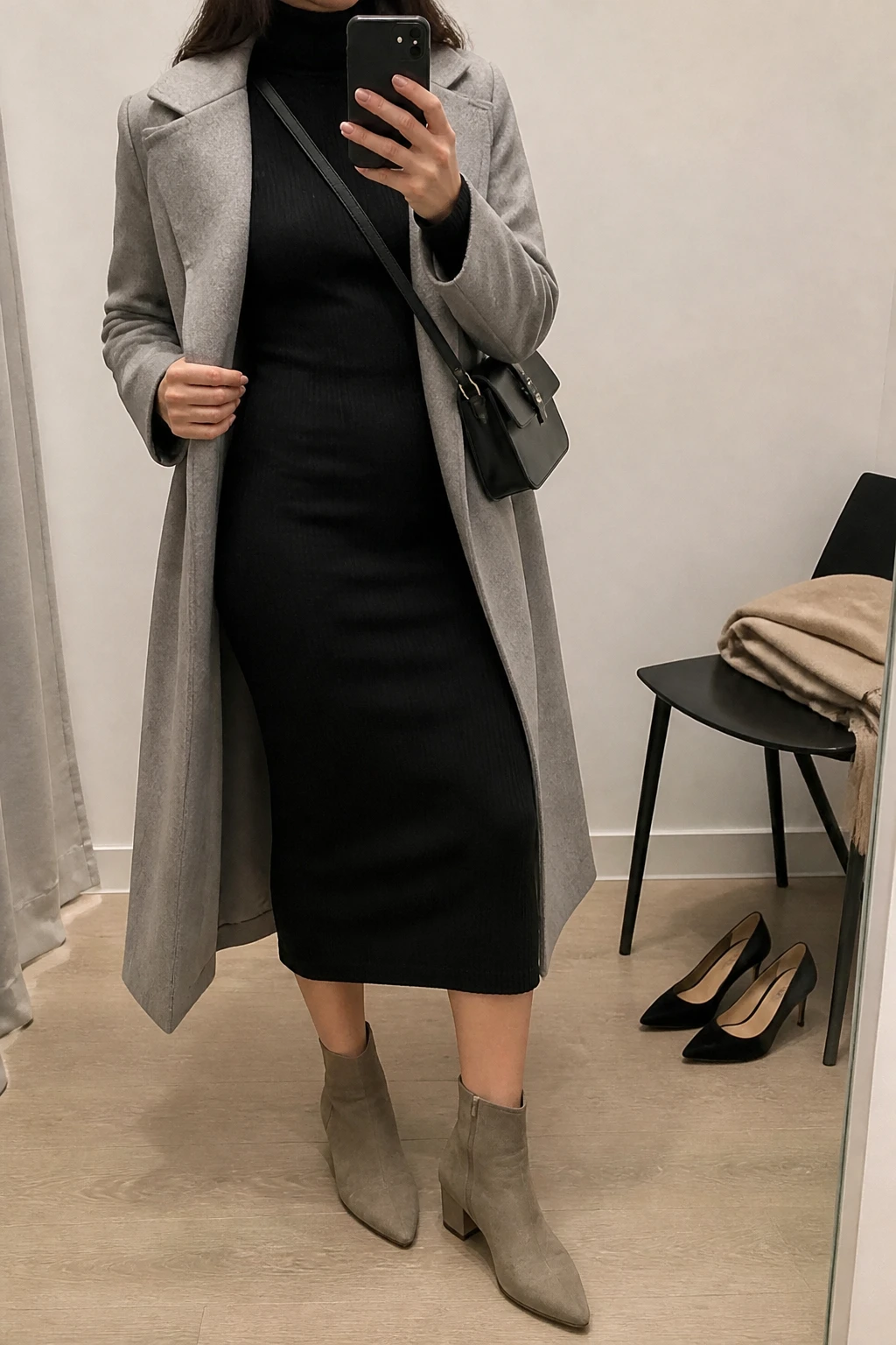 Image Prompt: Mirror selfie of a woman wearing a black long-sleeve fitted ribbed knit turtleneck midi dress, taupe suede pointed-toe ankle boots with low block heels, a long light grey wool coat worn open, a small black leather structured crossbody bag, and simple silver stud earrings, holding a phone in front of a mirror, cropped at the NECK so the face does not show, with a relaxed casual selfie pose, in a minimal dressing room, soft indoor lighting, a folded blanket on a chair and a pair of heels on the floor nearby, realistic ribbed knit fabric, soft suede texture and natural everyday pose, latest iPhone photo quality, no additional clothing or accessories.