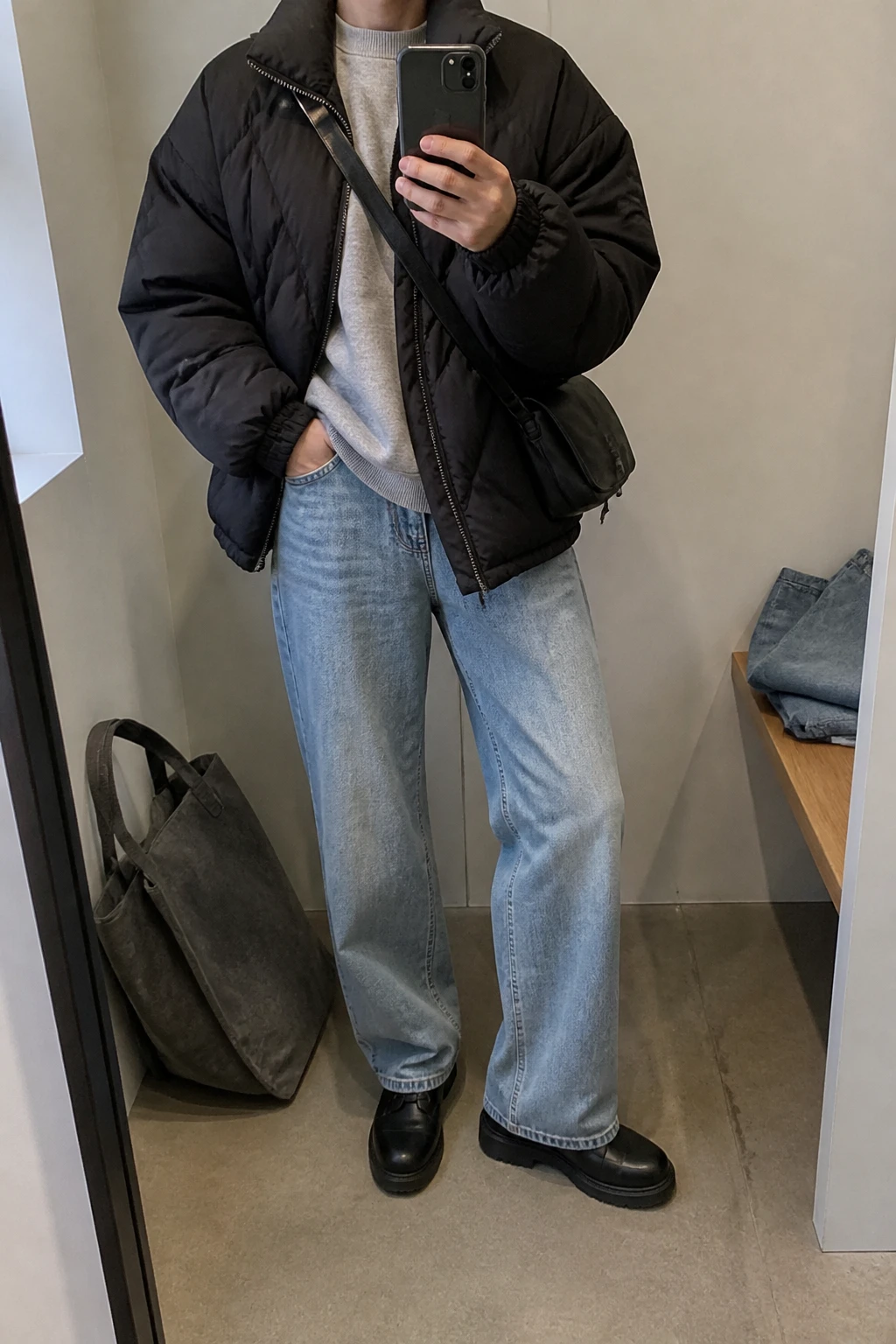 Image Prompt: Mirror selfie of a woman wearing a black oversized insulated quilted puffer jacket over a heather grey thick fleece-lined crewneck sweatshirt, light blue relaxed straight-leg jeans, black waterproof leather lace-up ankle boots with thick flat rubber soles, and a small black nylon crossbody bag, holding a phone in front of a mirror, cropped at the NECK so the face does not show, with a relaxed casual selfie pose, in a minimal dressing room, soft natural window light, a tote bag resting on the floor and folded denim jeans on a bench, realistic puffer jacket volume, natural light-wash denim creases and slightly imperfect casual styling, latest iPhone photo quality, no additional clothing or accessories.