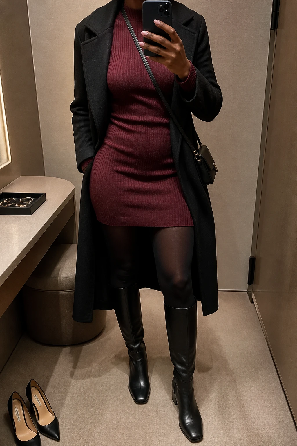 Image Prompt: Mirror selfie of a black woman wearing a burgundy long-sleeve fitted ribbed knit mini dress with a high neckline, black opaque thermal tights, black leather knee-high boots with low block heels, a long black wool-blend coat worn open, and a small black leather crossbody bag, holding a phone in front of a mirror, cropped at the NECK so the face does not show, with a relaxed casual selfie pose, in a minimal dressing room, warm moody lighting, a jewelry tray on the vanity and a pair of heels on the floor nearby, realistic ribbed knit texture, natural burgundy color and everyday fabric folds, latest iPhone photo quality, no additional clothing or accessories.