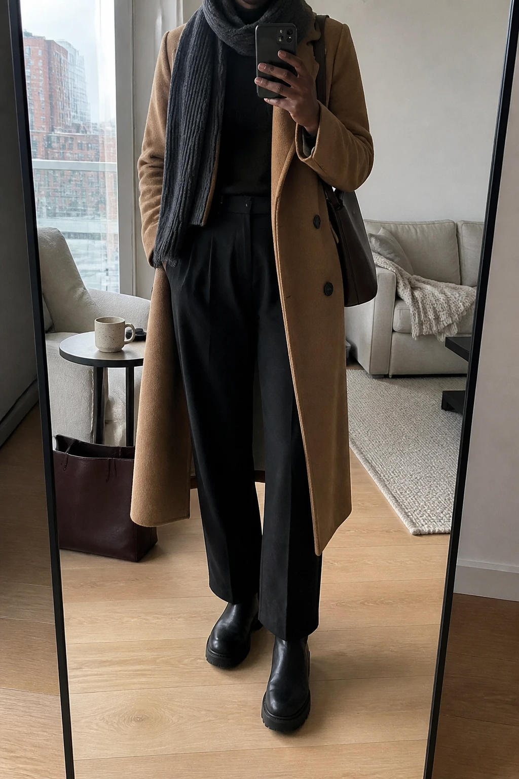 Image Prompt: Mirror selfie of a black woman wearing a long camel double-breasted wool coat over a black fitted cashmere turtleneck, black high-waisted wide-leg wool trousers, black leather chunky Chelsea boots, a structured dark brown leather shoulder bag, and a charcoal grey ribbed wool scarf wrapped loosely around the neck, holding a phone in front of a full-length mirror, cropped at the NECK so the face does not show, with a relaxed casual selfie pose, in a stylish city apartment, natural window light, a coffee mug on a side table and a tote bag resting on the floor, realistic fabric folds, natural proportions, slightly imperfect everyday appearance, latest iPhone photo quality, no additional clothing or accessories.