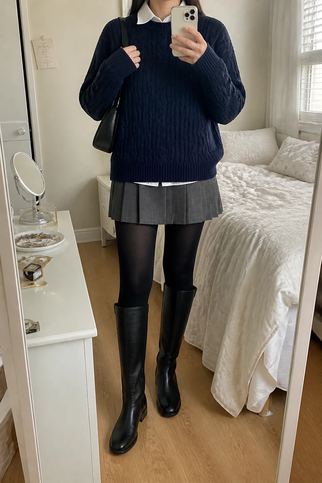 Image Prompt: Mirror selfie of a woman wearing a navy oversized cable-knit crewneck sweater layered over a white cotton collared button-front shirt with the collar visible, a charcoal grey wool-blend pleated mini skirt, black opaque fleece-lined tights, black leather knee-high flat boots with rubber soles, and a small black leather shoulder bag, holding a phone in front of a mirror, cropped at the NECK so the face does not show, with a relaxed casual selfie pose, in a feminine-themed bedroom, soft natural window light, soft pillows on an unmade bed and a jewelry tray on the vanity, realistic navy cable-knit stitches, natural grey skirt pleats, believable thermal tights, casual Korean-inspired styling, latest iPhone photo quality, no additional clothing or accessories.