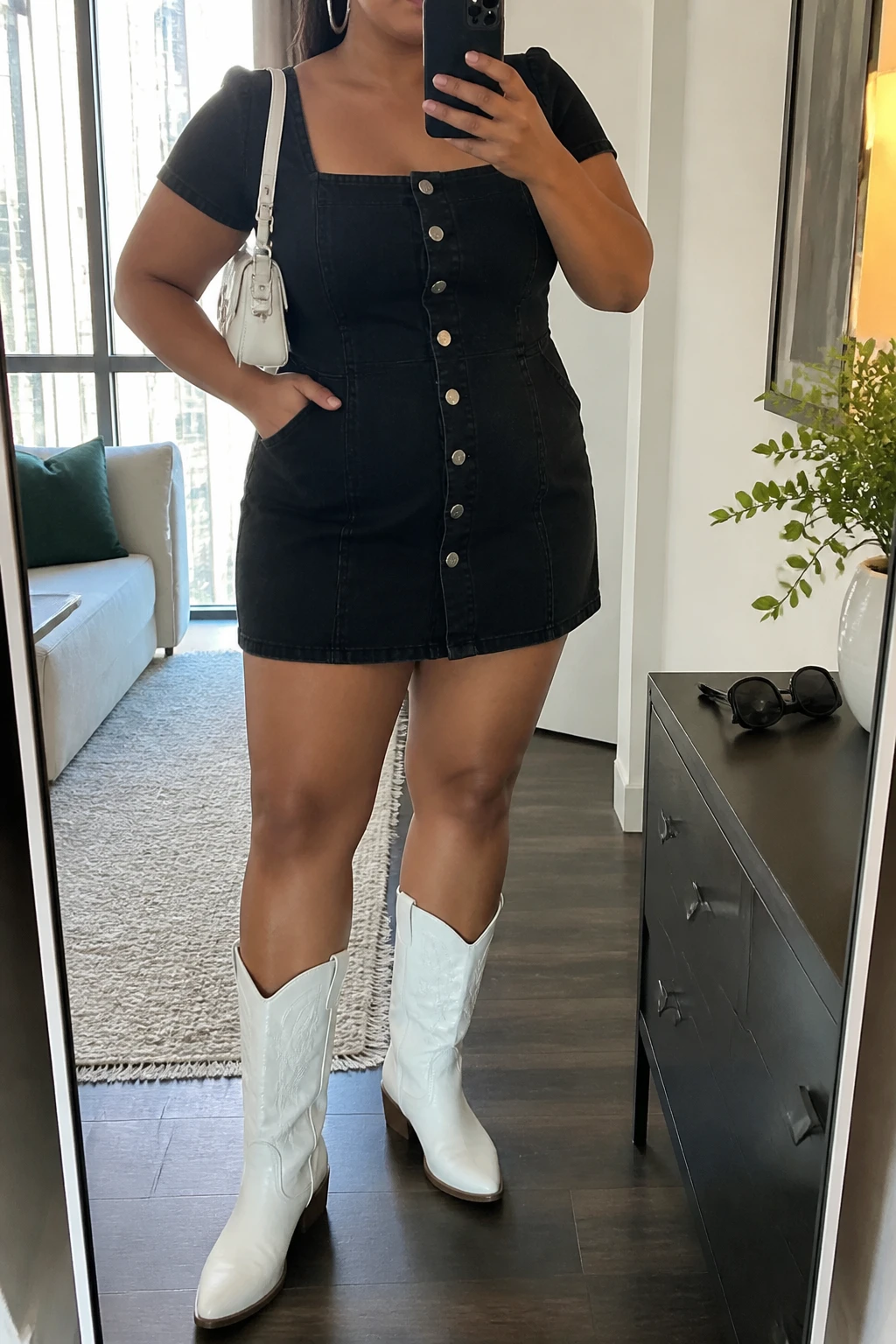 Mirror selfie of a plus size woman wearing a black denim mini dress with short sleeves, a square neckline, silver front buttons, and a softly fitted waist, white leather mid-calf cowboy boots with low stacked heels, a small white structured shoulder bag, and silver hoop earrings, clearly showing the exact clothing items, black denim, silver buttons, white footwear, and accessories listed, holding a phone in front of a mirror, cropped at the neck so the face does not show, relaxed confident selfie pose, in a stylish city apartment with natural window light, sunglasses placed on a dresser and a small plant nearby, natural unstaged appearance, latest iPhone photo quality.