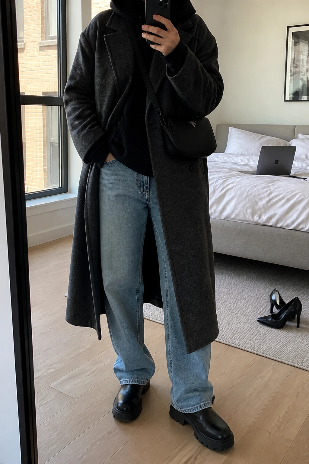 Image Prompt: Mirror selfie of a woman wearing a long charcoal grey oversized wool coat over a black thick fleece-lined hoodie, light blue baggy straight-leg denim jeans, black waterproof leather chunky Chelsea boots with flat lug soles, and a small black nylon crossbody bag, holding a phone in front of a mirror, cropped at the NECK so the face does not show, with a relaxed casual selfie pose, in a stylish city apartment, natural window light, a laptop on the bed and a pair of heels on the floor nearby, realistic charcoal wool coat texture, natural hoodie folds and relaxed denim shape, latest iPhone photo quality, no additional clothing or accessories.