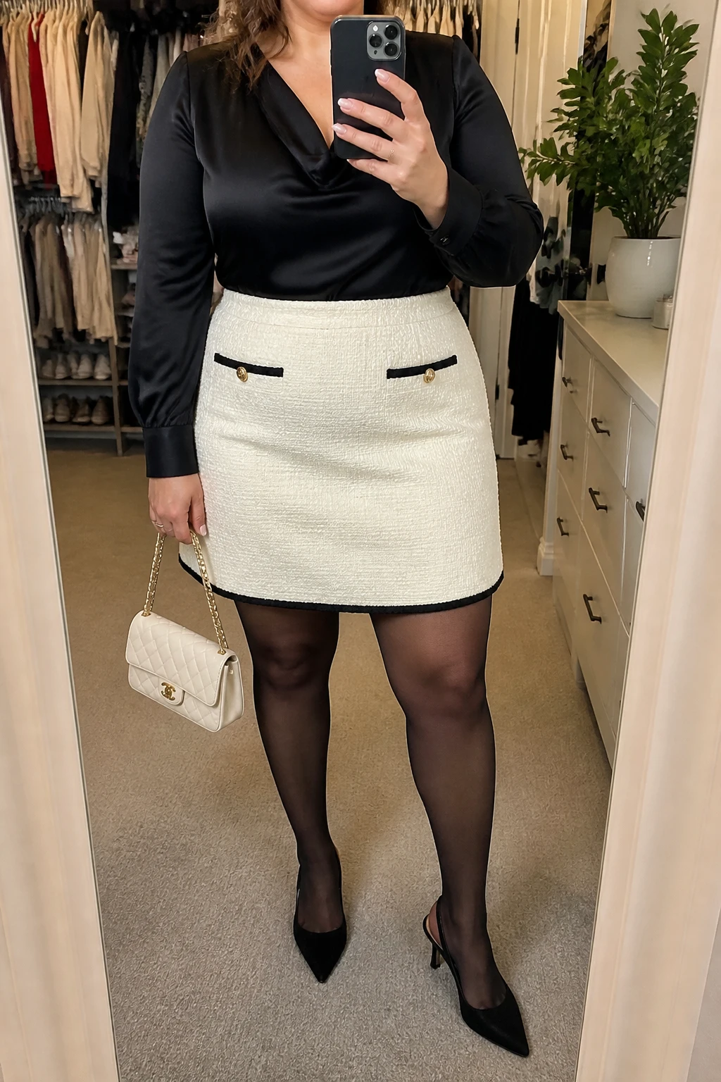 Mirror selfie of a plus size woman wearing a cream high-waisted bouclé mini skirt with black trim and a structured A-line shape, a black satin long-sleeve blouse with a softly draped neckline, black sheer tights, black pointed slingback heels, and a small cream quilted handbag, clearly showing the exact clothing items, cream bouclé, black trim, satin texture, footwear, and accessories listed, holding a phone in front of a mirror, cropped at the neck so her face does not appear, relaxed polished selfie stance, in a walk-in closet mirror with soft indoor lighting, an open closet with clothes visible and a small plant on a dresser, realistic elegant daytime outfit, latest iPhone photo quality.