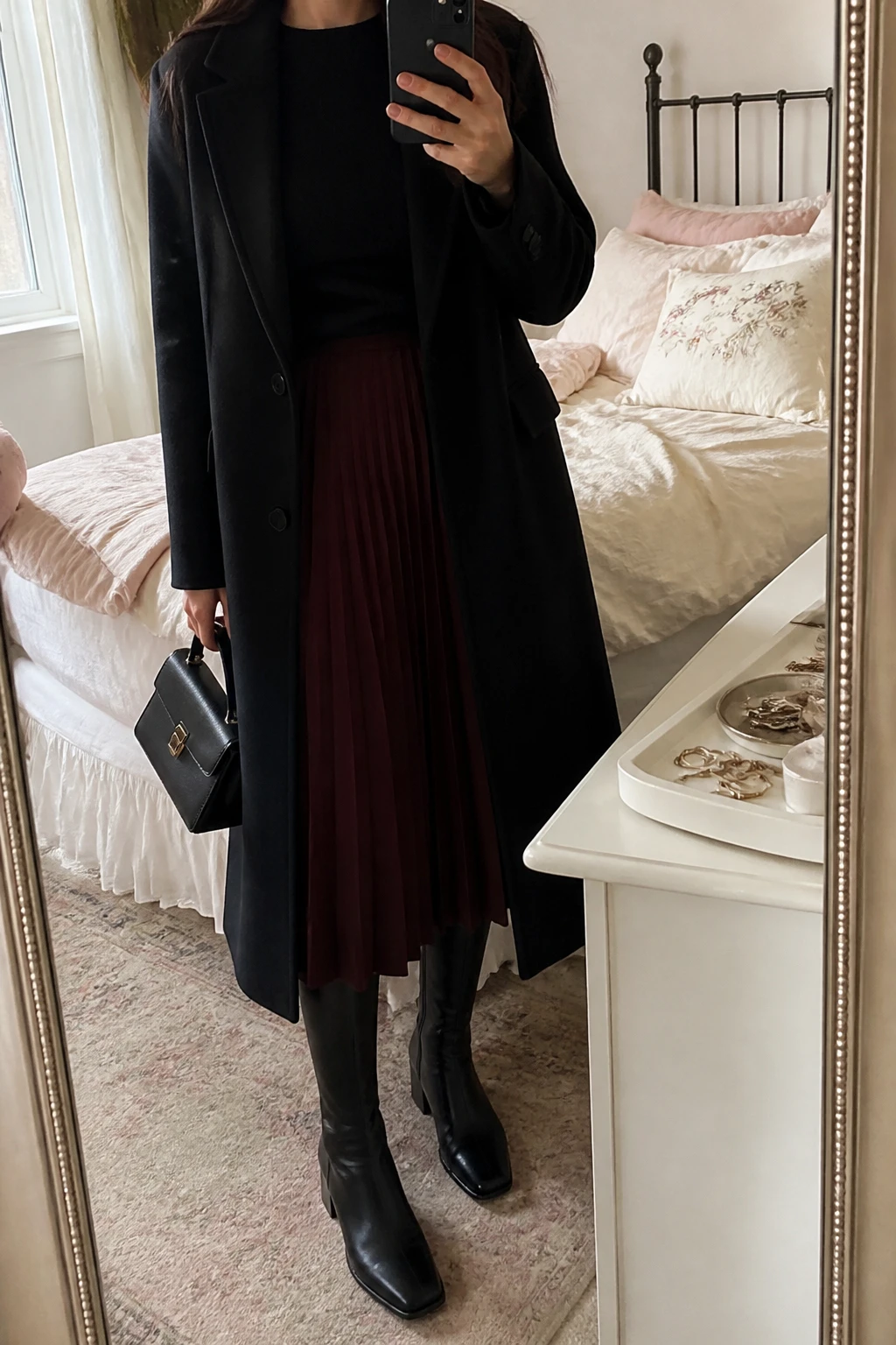 Image Prompt: Mirror selfie of a woman wearing a long black single-breasted tailored wool coat over a black fitted cashmere crewneck sweater tucked into a burgundy wool-blend pleated midi skirt, black leather knee-high boots with low block heels, a small structured black leather handbag, and black opaque thermal tights, holding a phone in front of a mirror, cropped at the NECK so the face does not show, with a relaxed casual selfie pose, in a feminine-themed bedroom, soft natural window light, soft pillows on an unmade bed and a jewelry tray on the vanity, realistic burgundy pleated wool fabric, natural black coat drape and understated Parisian styling, latest iPhone photo quality, no additional clothing or accessories.