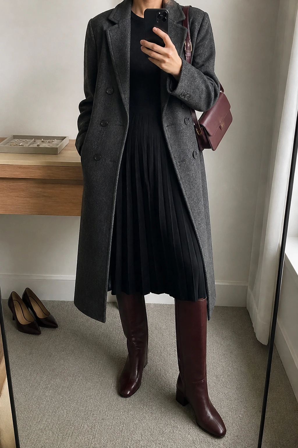Image Prompt: Mirror selfie of a woman wearing a black long-sleeve fine-knit midi dress with a softly pleated skirt and fitted waist, burgundy leather knee-high boots with low block heels, a long charcoal grey double-breasted wool coat worn open, a small burgundy leather shoulder bag, and delicate silver stud earrings, holding a phone in front of a mirror, cropped at the NECK so the face does not show, with a relaxed casual selfie pose, in a minimal dressing room, soft natural window light, a jewelry tray on the vanity and a pair of heels on the floor nearby, realistic black knit fabric, soft skirt pleats and deep burgundy leather texture, latest iPhone photo quality, no additional clothing or accessories.