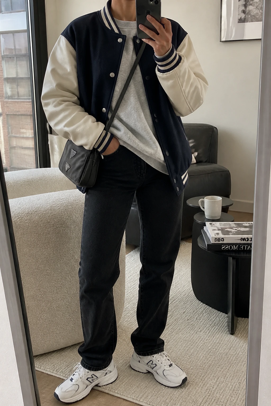 Image Prompt: Mirror selfie of a woman wearing a navy wool varsity jacket with cream faux-leather sleeves over a heather grey oversized fleece-lined crewneck sweatshirt, black high-waisted straight-leg denim jeans, white chunky leather sneakers with navy detailing, and a small charcoal grey nylon crossbody bag, holding a phone in front of a mirror, cropped at the NECK so the face does not show, with a relaxed casual selfie pose, in a stylish city apartment, soft natural window light, a coffee mug on a side table and a stack of fashion magazines on a side table, realistic navy varsity wool texture, natural grey fleece folds and black denim creases, latest iPhone photo quality, no additional clothing or accessories.