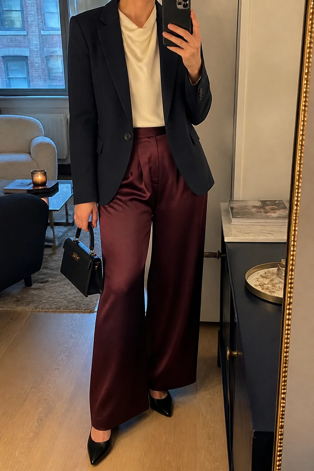 Image Prompt: Mirror selfie of a woman wearing a navy fitted single-breasted tailored wool blazer over an ivory silk blouse with a modest draped neckline, burgundy high-waisted wide-leg satin trousers, black leather pointed-toe block heels, a small black structured evening handbag, and delicate gold stud earrings, holding a phone in front of a mirror, cropped at the NECK so the face does not show, with a relaxed casual selfie pose, in a stylish city apartment, warm soft indoor lighting, a stack of fashion magazines on a side table and a jewelry tray on the vanity, realistic navy wool tailoring, natural burgundy satin trouser drape, understated evening colors, latest iPhone photo quality, no additional clothing or accessories.