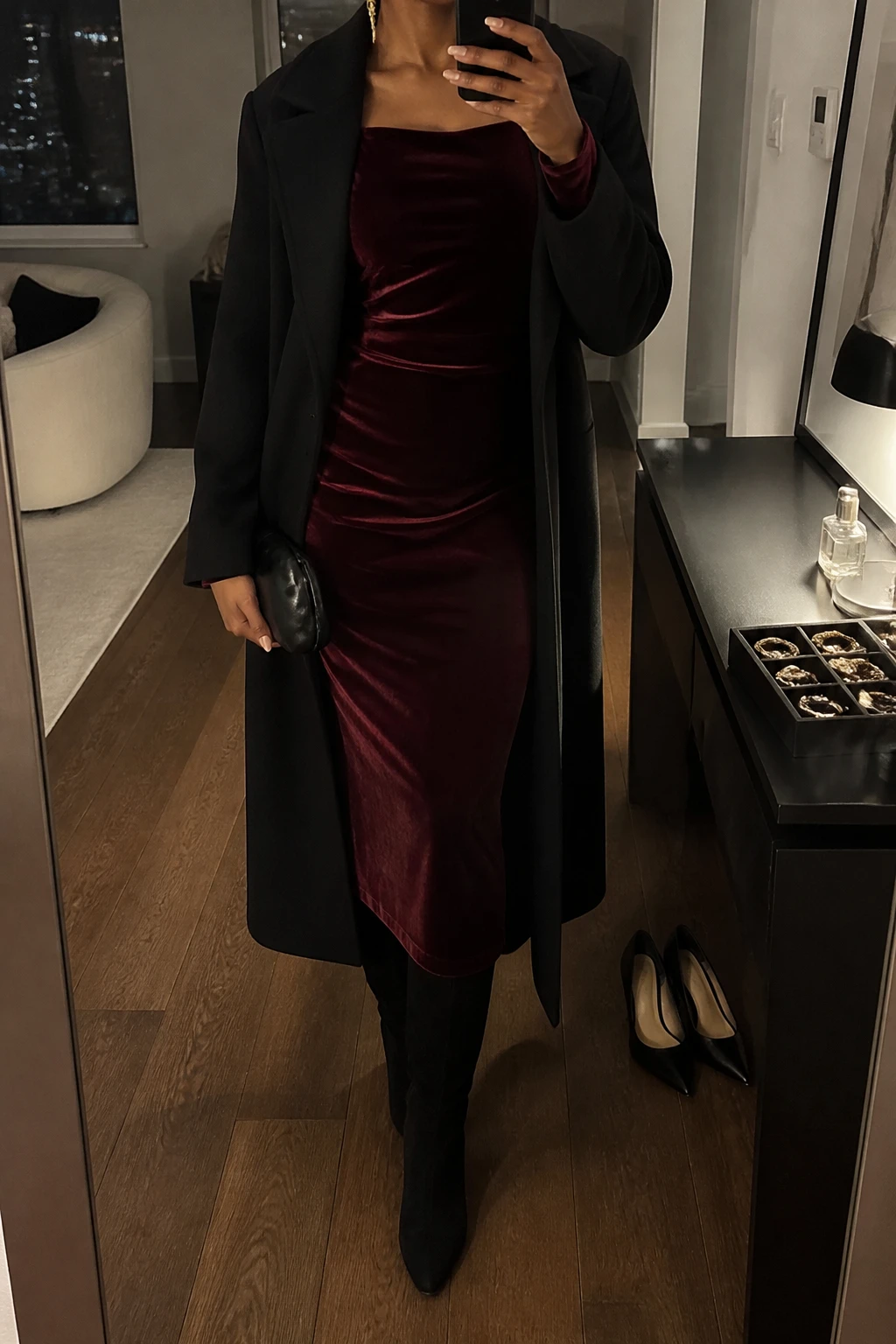 Image Prompt: Mirror selfie of a black woman wearing a burgundy long-sleeve velvet midi dress with a softly fitted waist and modest square neckline, black suede knee-high boots with low block heels, a long black tailored wool coat worn open, a small black satin clutch, and delicate gold drop earrings, holding a phone in front of a mirror, cropped at the NECK so the face does not show, with a relaxed casual selfie pose, in a stylish city apartment, warm moody indoor lighting, a jewelry tray on the vanity and a pair of heels on the floor nearby, realistic burgundy velvet texture, natural fabric drape and subtle warm shadows, latest iPhone photo quality, no additional clothing or accessories.