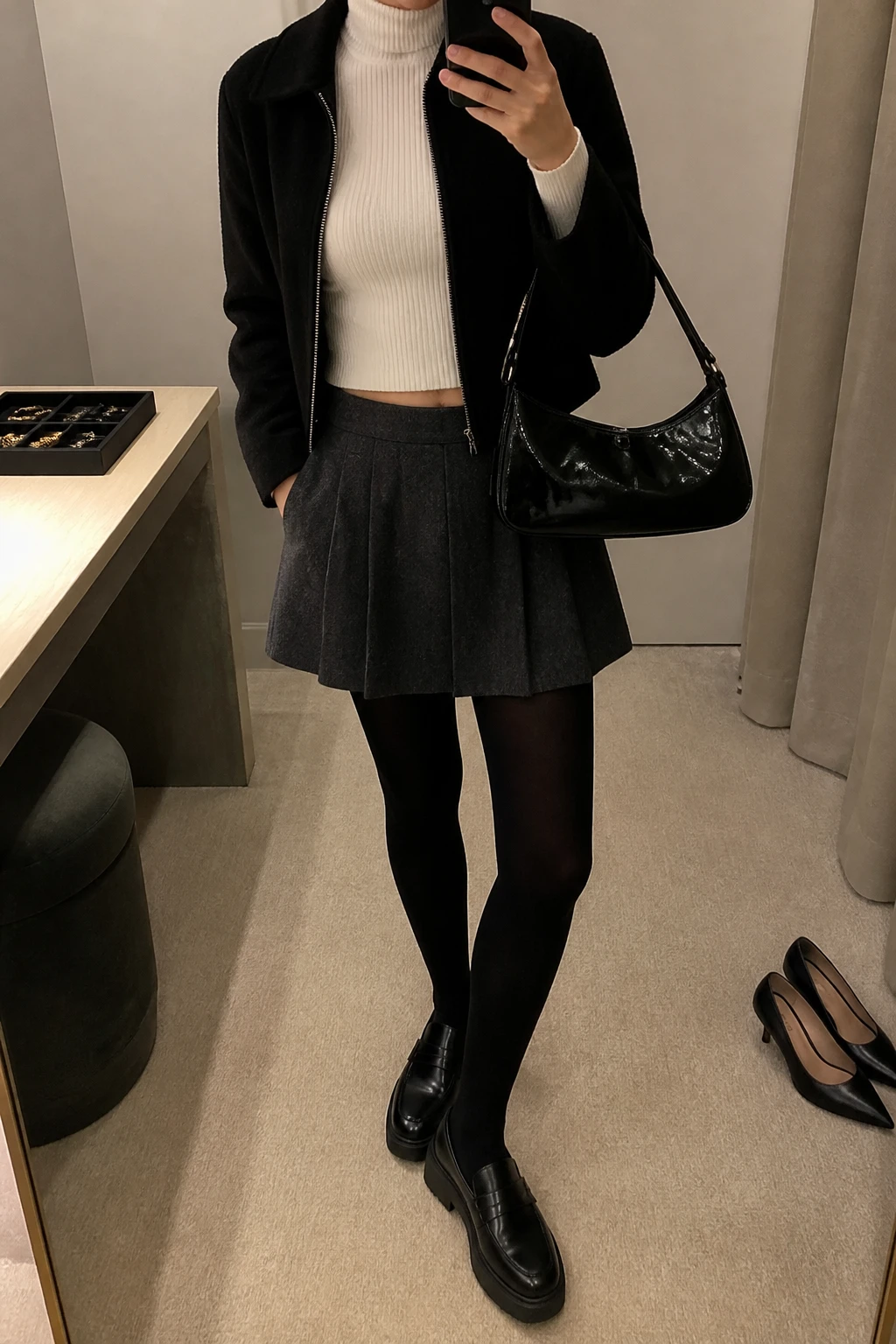Image Prompt: Mirror selfie of a woman wearing a cropped black wool-blend zip-front jacket over a white fitted ribbed turtleneck sweater, a charcoal grey pleated wool mini skirt, black opaque fleece-lined tights, black leather chunky platform loafers, and a small black patent leather baguette bag, holding a phone in front of a mirror, cropped at the NECK so the face does not show, with a relaxed casual selfie pose, in a minimal dressing room, warm soft indoor lighting, a jewelry tray on the vanity and a pair of heels on the floor nearby, realistic pleated wool fabric, natural black leather texture and subtle early 2000s preppy styling, latest iPhone photo quality, no additional clothing or accessories.