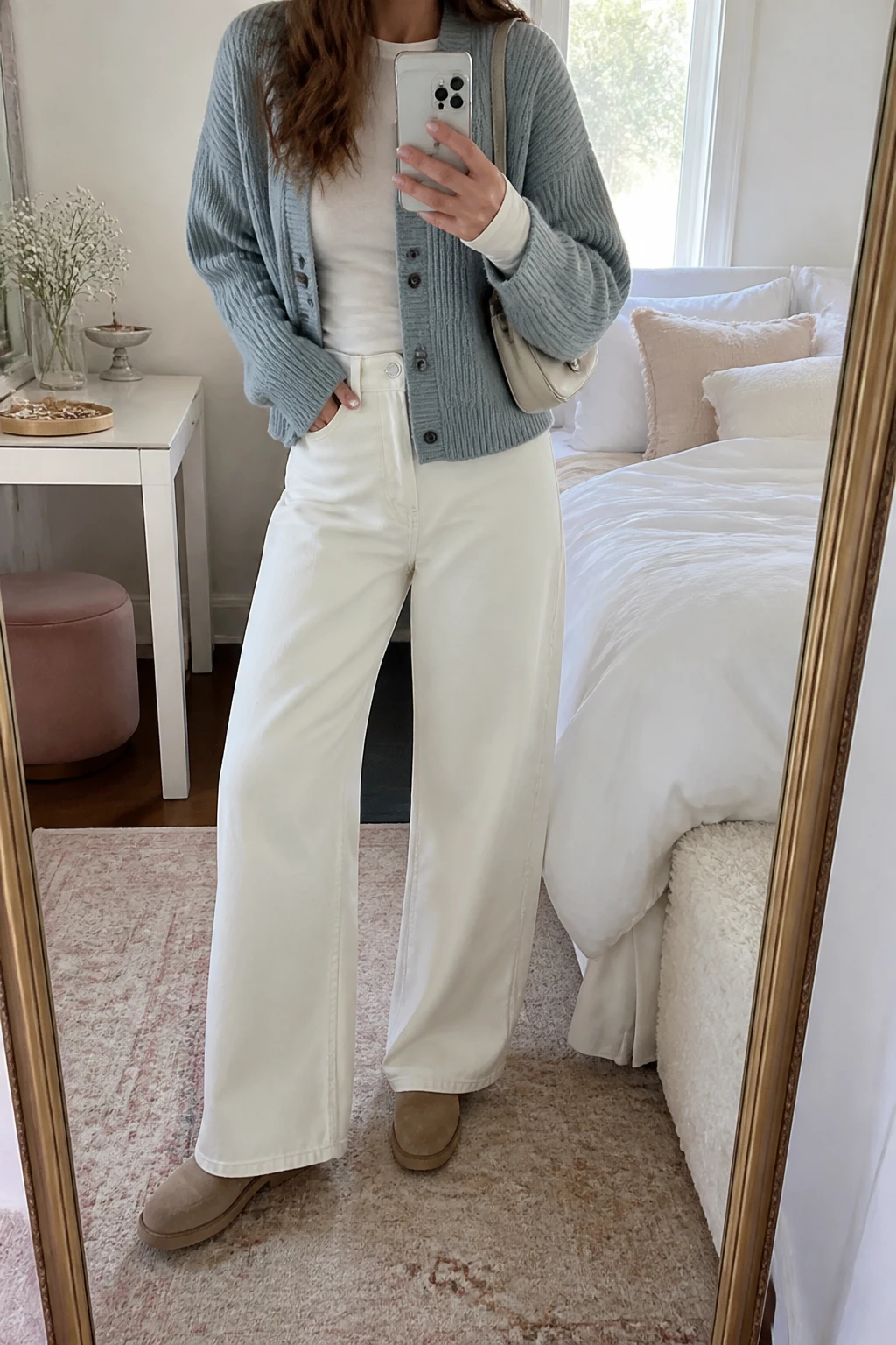 Image Prompt: Mirror selfie of a woman wearing a dusty blue chunky ribbed button-front cardigan over an ivory fitted long-sleeve cotton crewneck top, winter-white high-waisted wide-leg denim jeans, light taupe suede flat ankle boots, and a small cream leather shoulder bag, holding a phone in front of a mirror, cropped at the NECK so the face does not show, with a relaxed casual selfie pose, in a feminine-themed bedroom, soft natural window light, soft pillows on an unmade bed and a jewelry tray on the vanity, realistic dusty blue knit texture, natural white denim folds, soft pastel winter styling, latest iPhone photo quality, no additional clothing or accessories.