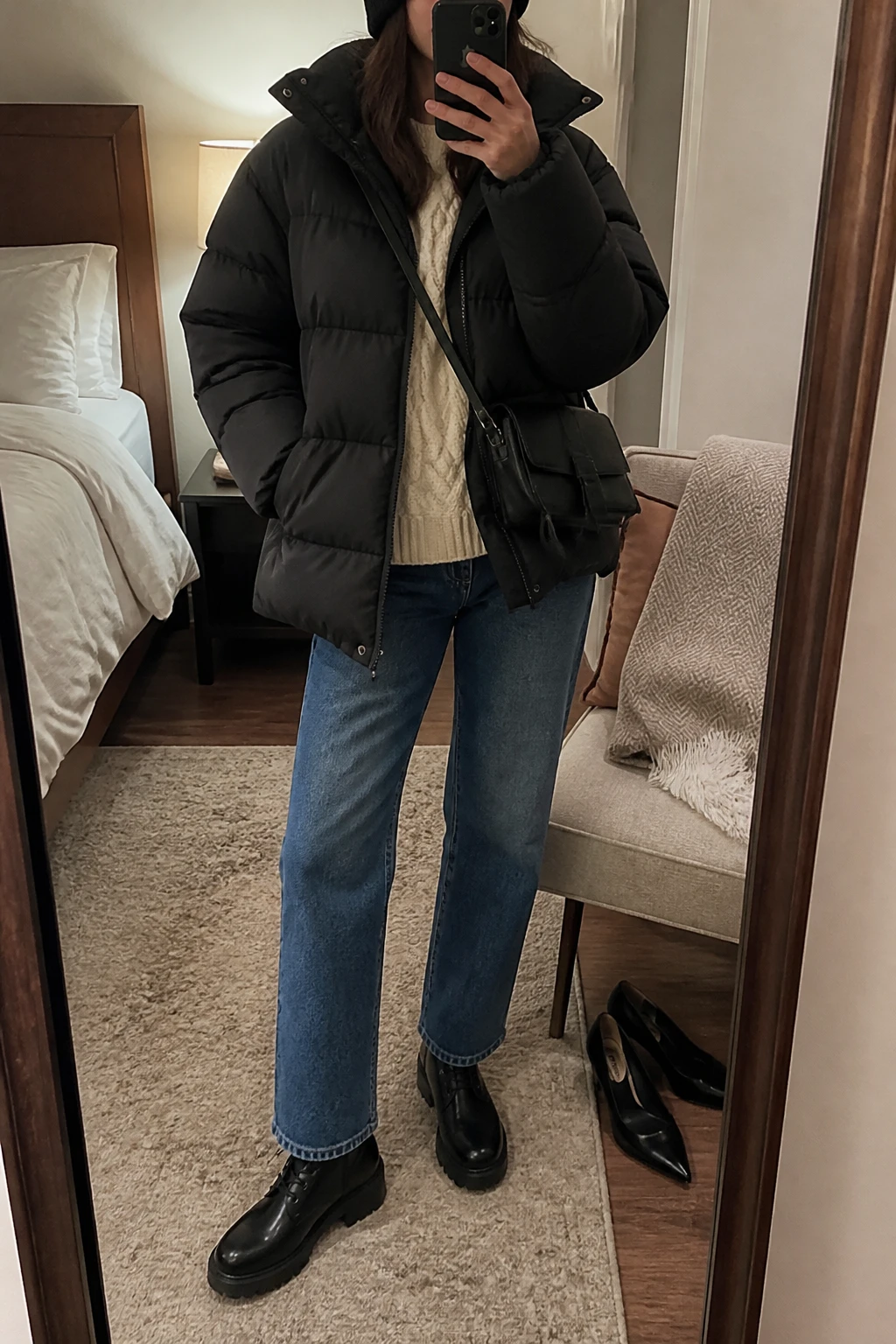 Image Prompt: Mirror selfie of a woman wearing a black oversized quilted down puffer jacket over a cream chunky cable-knit sweater, medium-wash blue straight-leg jeans, black waterproof leather lace-up ankle boots with thick rubber soles, a small black nylon crossbody bag, and a black ribbed knit beanie, holding a phone in front of a mirror, cropped at the NECK so the face does not show, with a relaxed casual selfie pose, in a cozy bedroom mirror setup, soft indoor lighting, a folded blanket on a chair and a pair of heels on the floor nearby, natural puffer volume, believable winter layering, latest iPhone photo quality, no additional clothing or accessories.