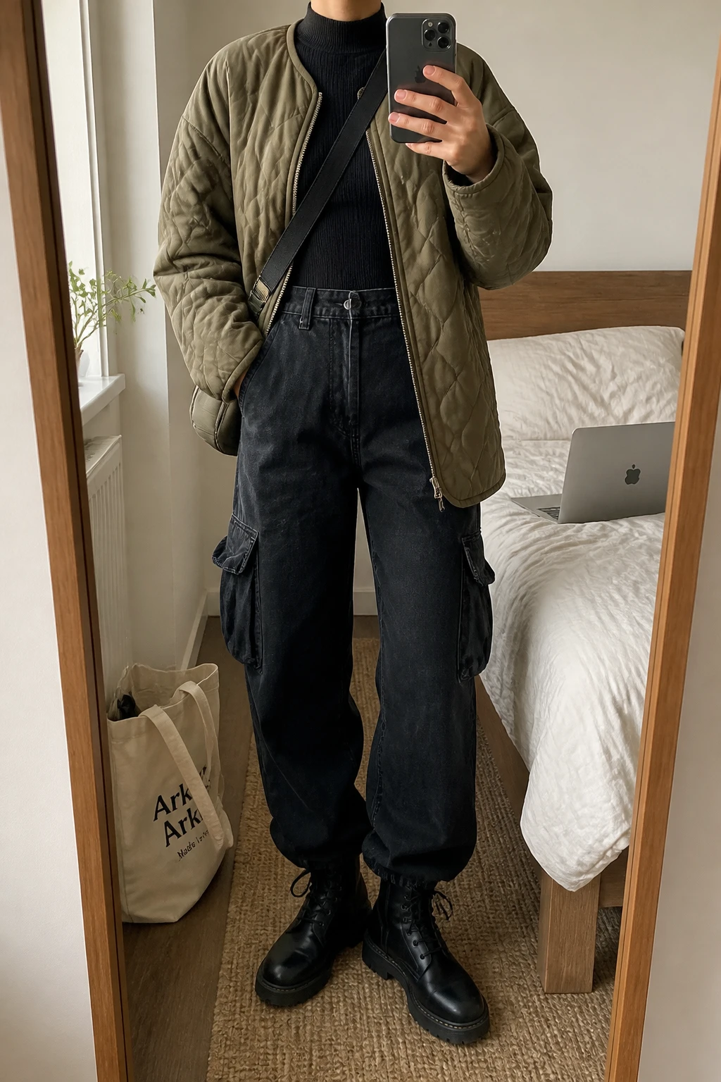 Image Prompt: Mirror selfie of a woman wearing an olive green oversized insulated quilted jacket over a black fitted thermal mock-neck top, black high-waisted baggy denim cargo jeans with large side pockets, black leather chunky lace-up boots with flat rubber soles, and a small dark olive nylon crossbody bag, holding a phone in front of a mirror, cropped at the NECK so the face does not show, with a relaxed casual selfie pose, in a cozy bedroom mirror setup, soft natural window light, a laptop on the bed and a tote bag resting on the floor, realistic olive quilted fabric, natural black cargo denim folds and casual streetwear styling, latest iPhone photo quality, no additional clothing or accessories.
