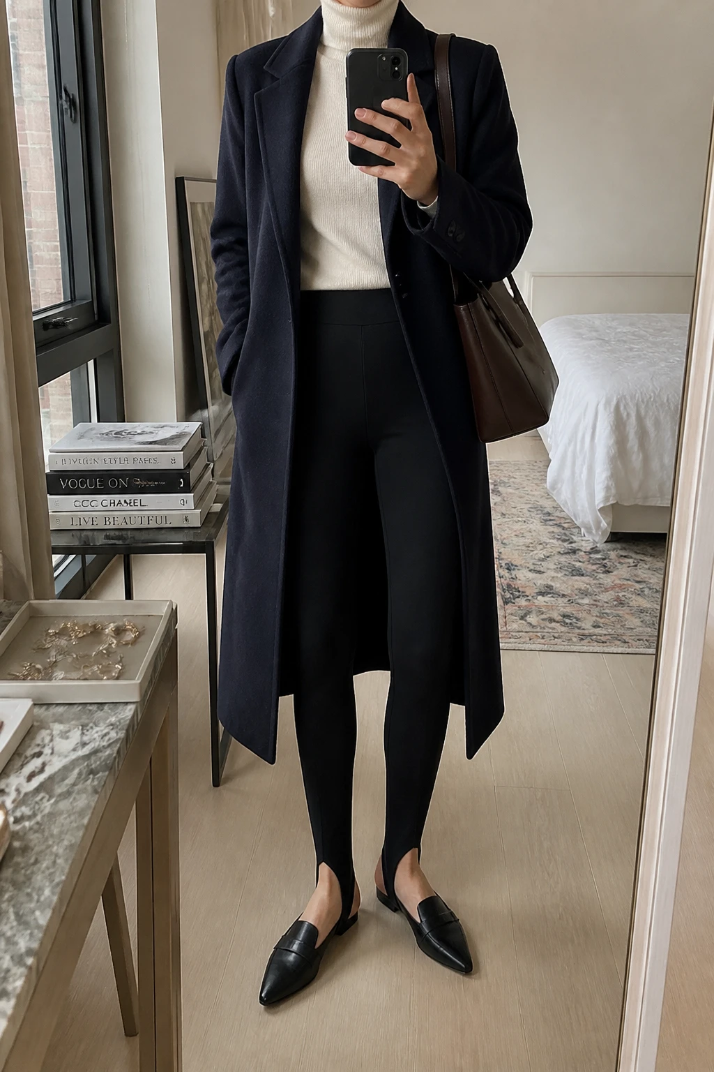 Image Prompt: Mirror selfie of a woman wearing a long navy tailored wool coat over a cream fitted cashmere turtleneck sweater, black high-waisted thick ponte-knit stirrup leggings with the stirrup straps tucked beneath the feet, black leather pointed-toe flat loafers, and a structured dark brown leather shoulder bag, holding a phone in front of a mirror, cropped at the NECK so the face does not show, with a relaxed casual selfie pose, in a stylish city apartment, soft natural window light, a stack of fashion magazines on a side table and a jewelry tray on the vanity, realistic navy wool texture, natural black ponte fabric and polished everyday styling, latest iPhone photo quality, no additional clothing or accessories.