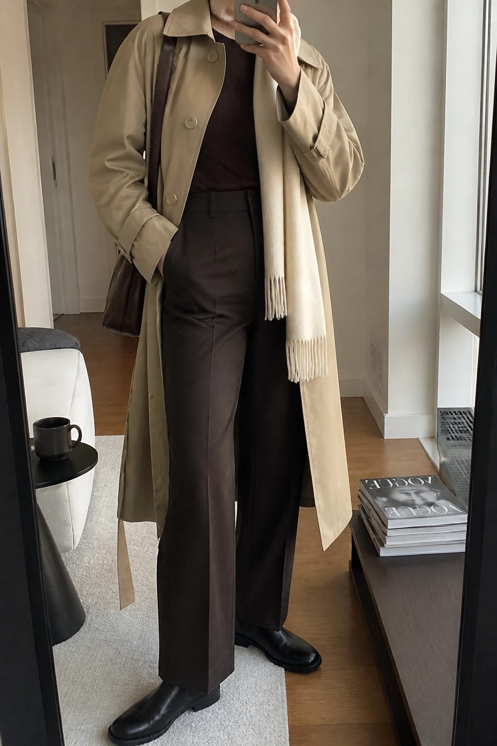 Image Prompt: Mirror selfie of a woman wearing a long beige water-resistant belted trench coat over a chocolate brown fitted merino wool crewneck sweater, dark chocolate high-waisted straight-leg tailored trousers, black leather flat Chelsea boots with rubber soles, a structured dark brown leather shoulder bag, and a cream wool scarf, holding a phone in front of a mirror, cropped at the NECK so the face does not show, with a relaxed casual selfie pose, in a stylish city apartment, soft natural window light, a coffee mug on a side table and a stack of fashion magazines on a side table, realistic beige trench fabric, natural chocolate trouser creases and soft cream wool texture, latest iPhone photo quality, no additional clothing or accessories.