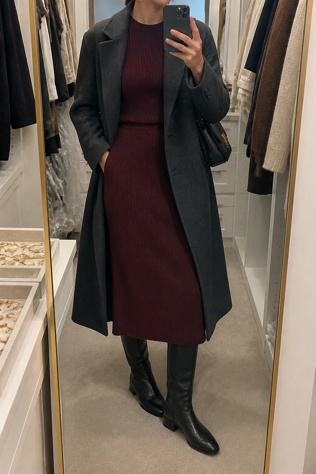 Image Prompt: Mirror selfie of a woman wearing a burgundy long-sleeve ribbed wool-blend midi dress with a fitted waist, black leather knee-high boots with low block heels, a long charcoal grey tailored wool coat worn open, a structured black leather shoulder bag, and delicate gold hoop earrings, holding a phone in front of a mirror, cropped at the NECK so the face does not show, with a relaxed casual selfie pose, in a walk-in closet mirror, soft indoor lighting, an open closet with clothes visible and a jewelry tray on the vanity, realistic burgundy knit texture and natural winter fabric folds, latest iPhone photo quality, no additional clothing or accessories.