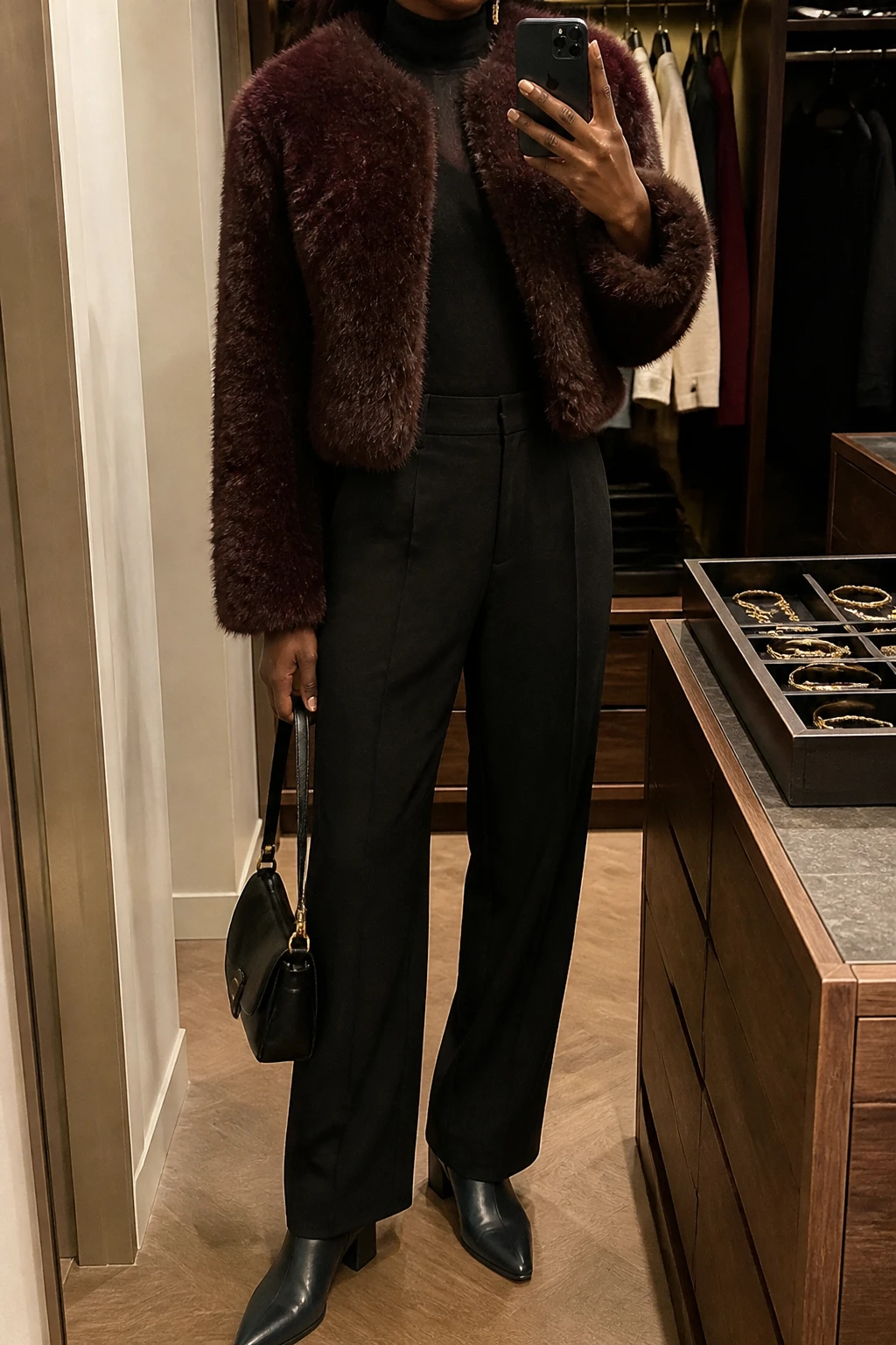 Image Prompt: Mirror selfie of a black woman wearing a cropped burgundy faux-fur jacket over a black fitted fine-knit turtleneck sweater, black high-waisted straight-leg tailored wool trousers, black leather pointed-toe low block-heel ankle boots, a small black structured leather handbag, and delicate gold drop earrings, holding a phone in front of a mirror, cropped at the NECK so the face does not show, with a relaxed casual selfie pose, in a walk-in closet mirror, warm moody indoor lighting, an open closet with clothes visible and a jewelry tray on the vanity, realistic burgundy faux-fur texture, natural black trouser drape, believable rich brown skin visible on hands, latest iPhone photo quality, no additional clothing or accessories.