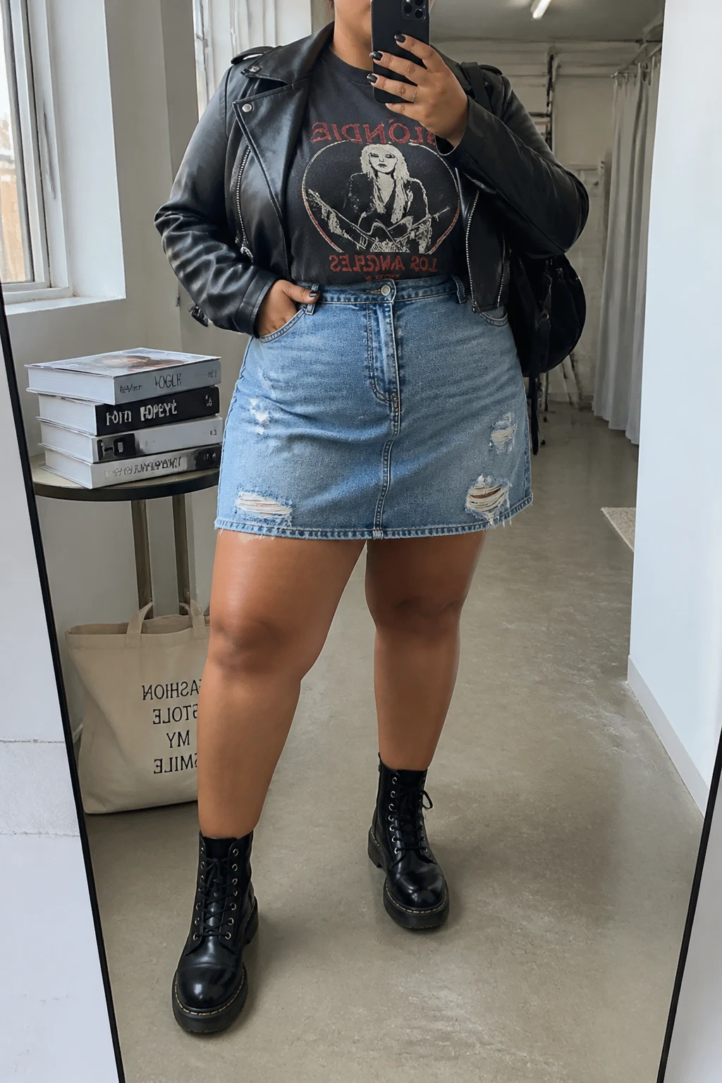 Mirror selfie of a plus size woman wearing a medium-wash distressed denim mini skirt with a relaxed A-line shape, a fitted faded black graphic T-shirt tucked into the skirt, a cropped black leather jacket worn open, black lace-up combat boots, and a small black nylon backpack, clearly showing the exact clothing items, denim distressing, graphic print, black jacket, footwear, and accessories listed, holding a phone in front of a mirror, cropped at the neck so her face is hidden, relaxed edgy selfie pose, in a fashion studio mirror corner with natural window light, a stack of fashion magazines on a side table and a tote bag resting on the floor, realistic casual concert outfit, latest iPhone photo quality.