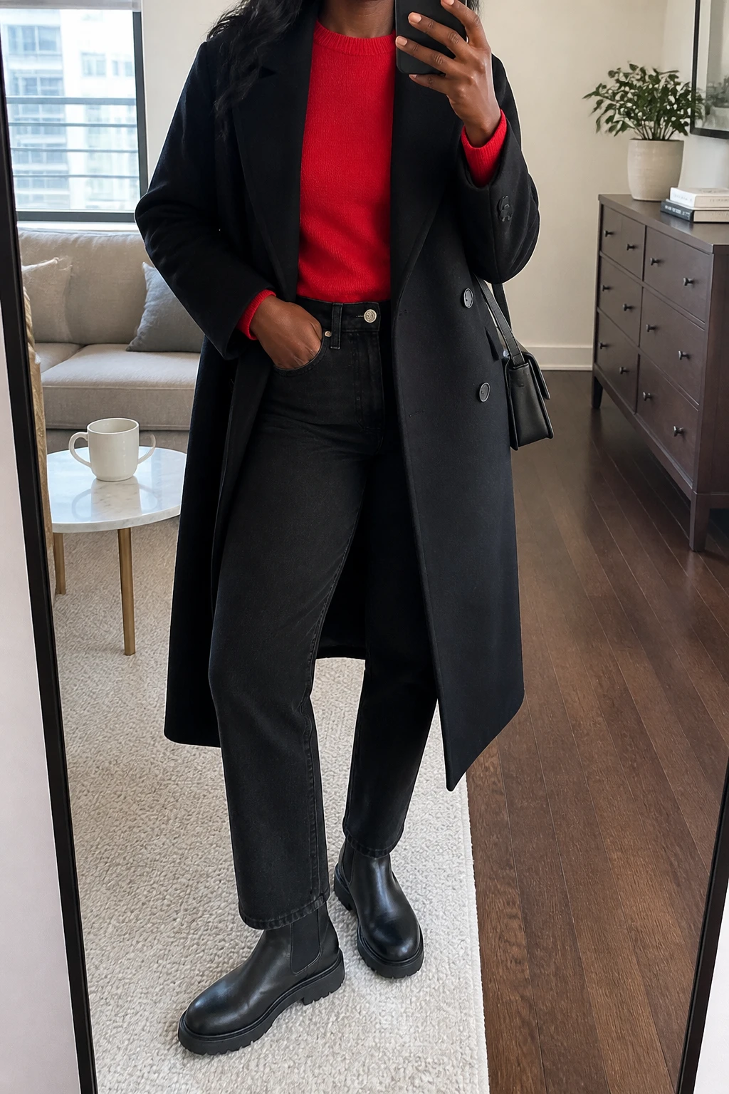Image Prompt: Mirror selfie of a black woman wearing a long black double-breasted tailored wool coat worn open over a bright cool cherry red fitted cashmere crewneck sweater, black high-waisted straight-leg denim jeans, black leather chunky Chelsea boots with flat rubber soles, a small black structured shoulder bag, and delicate silver stud earrings, holding a phone in front of a mirror, cropped at the NECK so the face does not show, with a relaxed casual selfie pose, in a stylish city apartment, soft natural window light, a coffee mug on a side table and a small plant on a dresser, realistic bright red cashmere knit texture, natural black denim folds, clear cool red color without oversaturation, latest iPhone photo quality, no additional clothing or accessories.