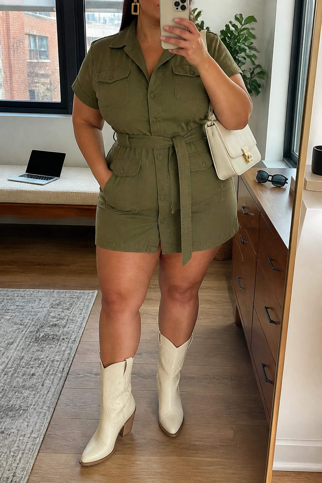 Mirror selfie of a plus size woman wearing an olive green utility mini dress with short sleeves, a button front, flat chest pockets, and a matching fabric belt, cream leather mid-calf cowboy boots, a cream structured shoulder bag, and gold rectangular earrings, clearly showing the exact dress, olive fabric, utility pockets, belt, cream boots, and accessories listed, holding a phone in front of a mirror, cropped at the neck so the face does not show, relaxed confident selfie pose, in a stylish city apartment with natural window light, a laptop on a bench and sunglasses placed on a dresser, realistic modern Western styling, latest iPhone photo quality.