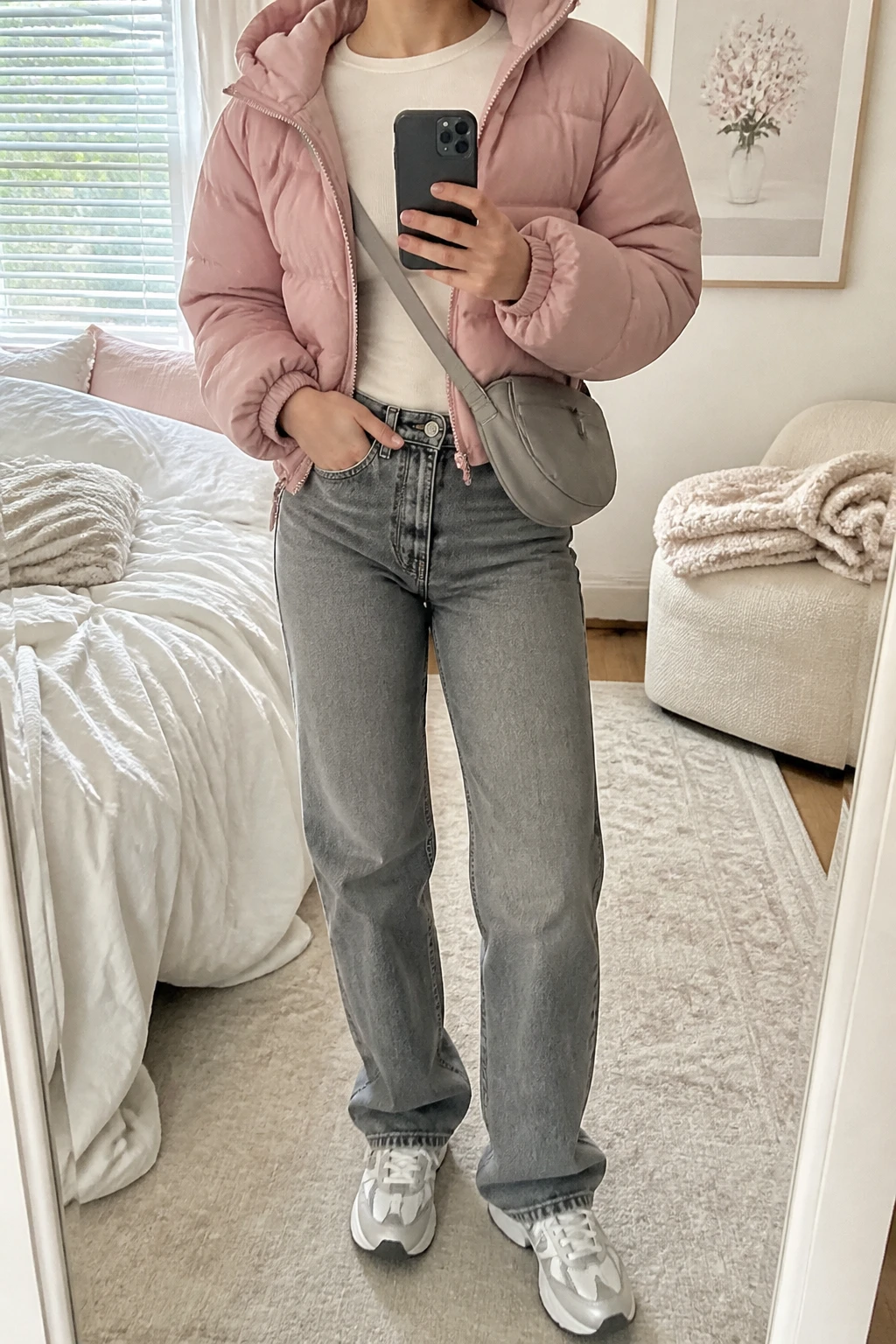 Image Prompt: Mirror selfie of a woman wearing a dusty pink insulated hooded cropped puffer jacket over an ivory fitted thermal crewneck top, washed grey high-waisted straight-leg denim jeans, white chunky leather sneakers with light grey detailing, and a small dove grey nylon crossbody bag, holding a phone in front of a mirror, cropped at the NECK so the face does not show, with a relaxed casual selfie pose, in a feminine-themed bedroom, soft natural window light, soft pillows on an unmade bed and a folded blanket on a chair, realistic dusty pink quilted fabric, natural grey denim creases and soft everyday lighting, latest iPhone photo quality, no additional clothing or accessories.