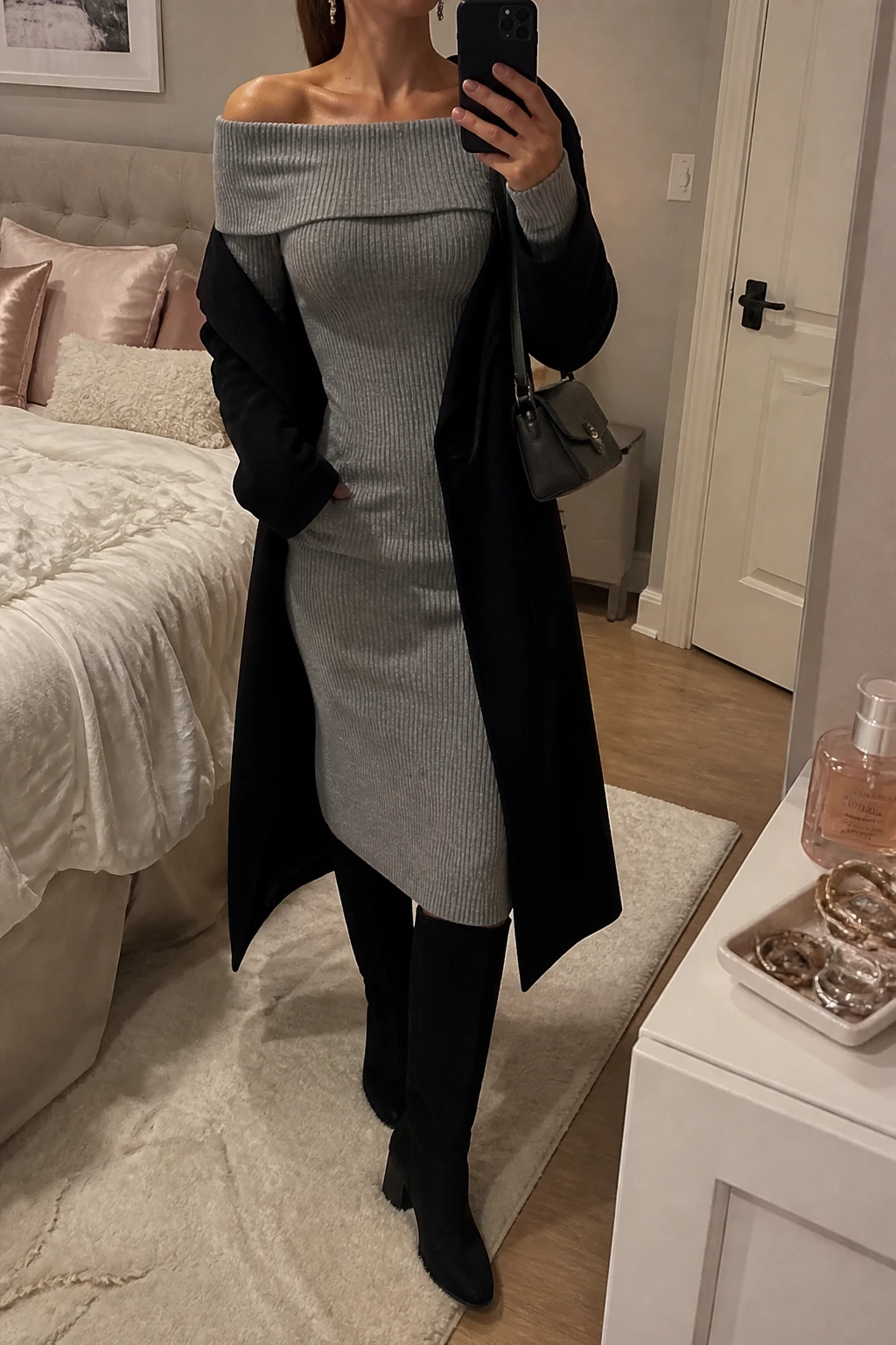 Image Prompt: Mirror selfie of a woman wearing a heather grey long-sleeve off-shoulder ribbed knit midi dress with a folded neckline and softly fitted waist, black suede knee-high boots with low block heels, a long black wool wrap coat worn open, a small charcoal grey leather shoulder bag, and delicate silver drop earrings, holding a phone in front of a mirror, cropped at the NECK so the face does not show, with a relaxed casual selfie pose, in a feminine-themed bedroom, warm soft indoor lighting, soft pillows on an unmade bed and a jewelry tray on the vanity, realistic grey ribbed knit texture, natural black suede finish and subtle evening shadows, latest iPhone photo quality, no additional clothing or accessories.