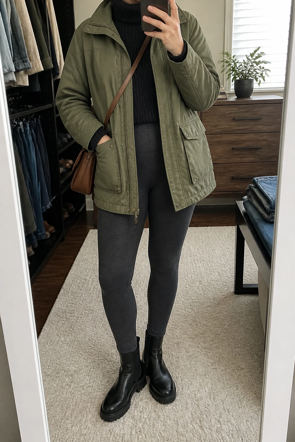 Image Prompt: Mirror selfie of a woman wearing an olive green insulated utility jacket with a relaxed fit and large front pockets over a black chunky ribbed turtleneck sweater, charcoal grey high-waisted thermal leggings, black leather chunky flat Chelsea boots, and a medium dark brown leather crossbody bag, holding a phone in front of a mirror, cropped at the NECK so the face does not show, with a relaxed casual selfie pose, in a walk-in closet mirror, soft natural window light, folded denim jeans on a bench and a small plant on a dresser, realistic olive jacket fabric, natural charcoal grey leggings texture and casual winter proportions, latest iPhone photo quality, no additional clothing or accessories.
