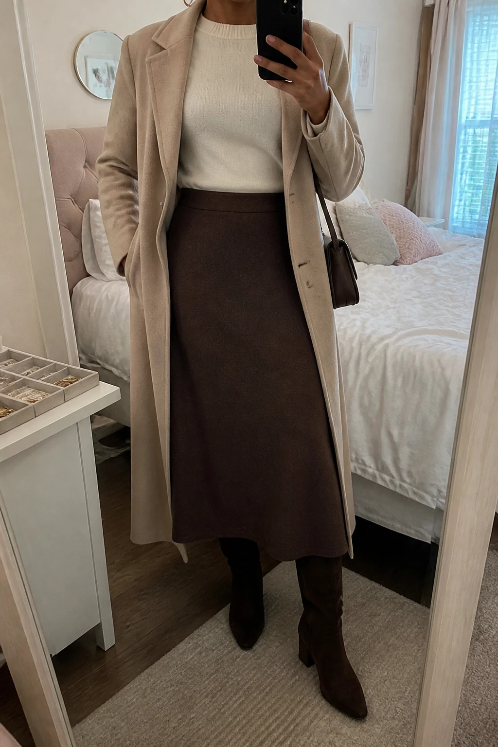 Image Prompt: Mirror selfie of a black woman wearing a cream fitted cashmere crewneck sweater tucked into a chocolate brown wool-blend A-line midi skirt, dark brown suede knee-high boots with low block heels, a warm beige tailored wool coat worn open, a small structured dark brown leather shoulder bag, and delicate gold hoop earrings, holding a phone in front of a mirror, cropped at the NECK so the face does not show, with a relaxed casual selfie pose, in a feminine-themed bedroom, soft natural window light, a jewelry tray on the vanity and soft pillows on an unmade bed, realistic cream cashmere and brown wool textures, latest iPhone photo quality, no additional clothing or accessories.