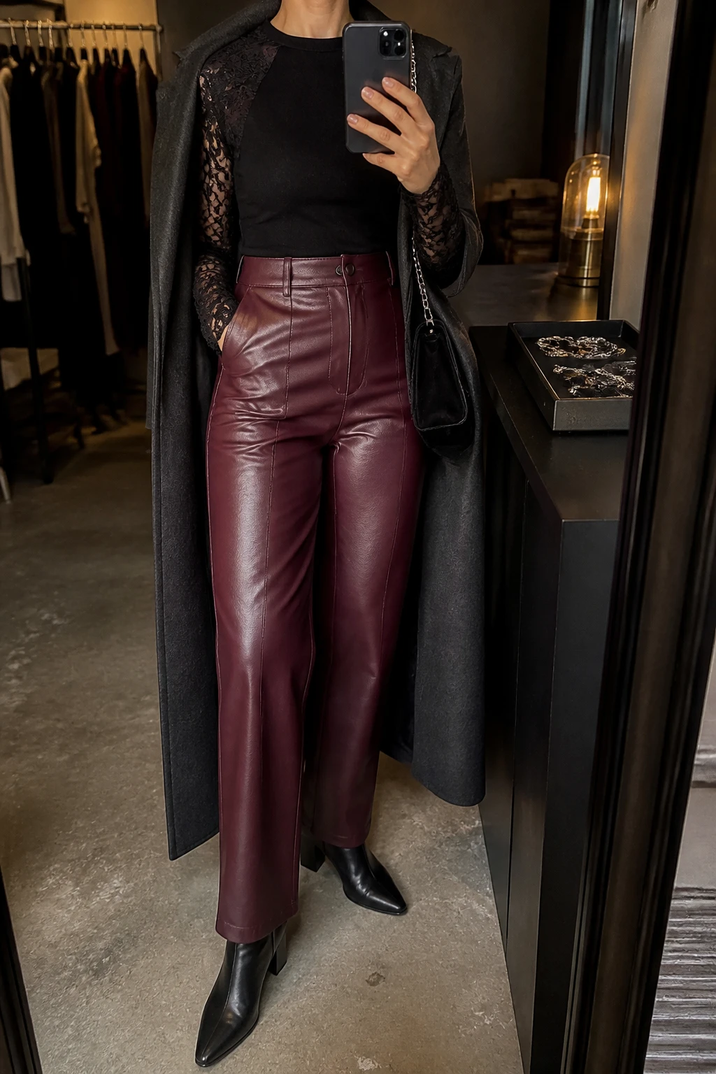 Image Prompt: Mirror selfie of a woman wearing a black fitted long-sleeve top with a solid black bodice and delicate semi-sheer floral lace sleeves, deep burgundy high-waisted straight-leg faux-leather trousers, black leather pointed-toe ankle boots with block heels, a long charcoal grey tailored wool coat worn open, a small black velvet shoulder bag, and delicate silver oval earrings, holding a phone in front of a mirror, cropped at the NECK so the face does not show, with a relaxed casual selfie pose, in a fashion studio mirror corner, warm moody lighting, a clothing rack with garments behind and a jewelry tray on the vanity, realistic burgundy leather grain, natural black floral lace detail and subtle evening shadows, latest iPhone photo quality, no additional clothing or accessories.