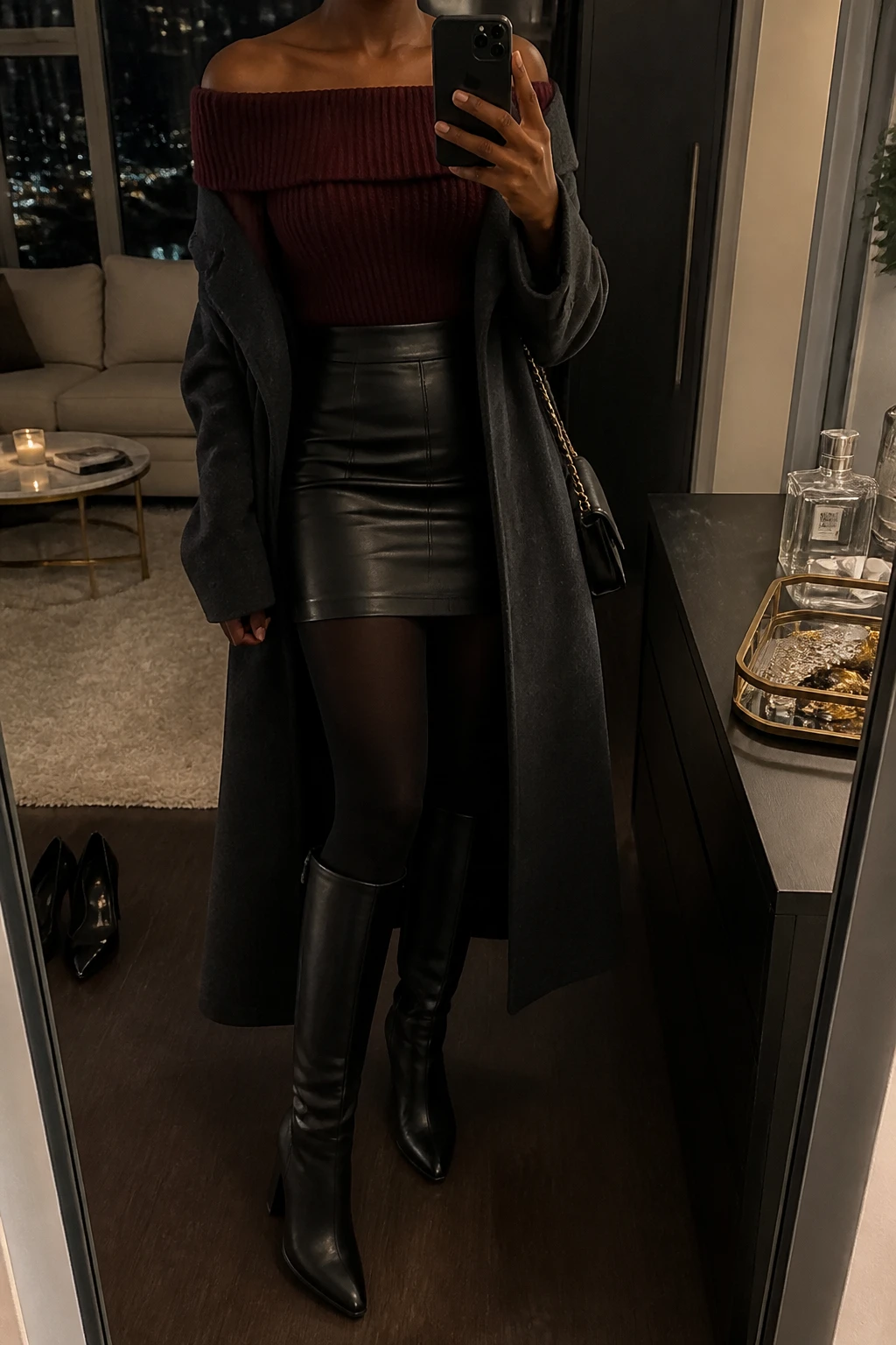 Image Prompt: Mirror selfie of a black woman wearing a deep burgundy fitted off-shoulder chunky-knit sweater with a folded neckline, a black high-waisted faux-leather mini skirt, black opaque fleece-lined tights, black leather knee-high boots with block heels, a long charcoal grey tailored wool coat worn open, and a small black chain-strap shoulder bag, holding a phone in front of a full-length mirror, cropped at the NECK so the face does not show, with a relaxed casual selfie pose, in a stylish city apartment, warm moody lighting, a jewelry tray on the vanity and a pair of heels on the floor, realistic burgundy knit texture, natural black leather grain, soft evening shadows, latest iPhone photo quality, no additional clothing or accessories.