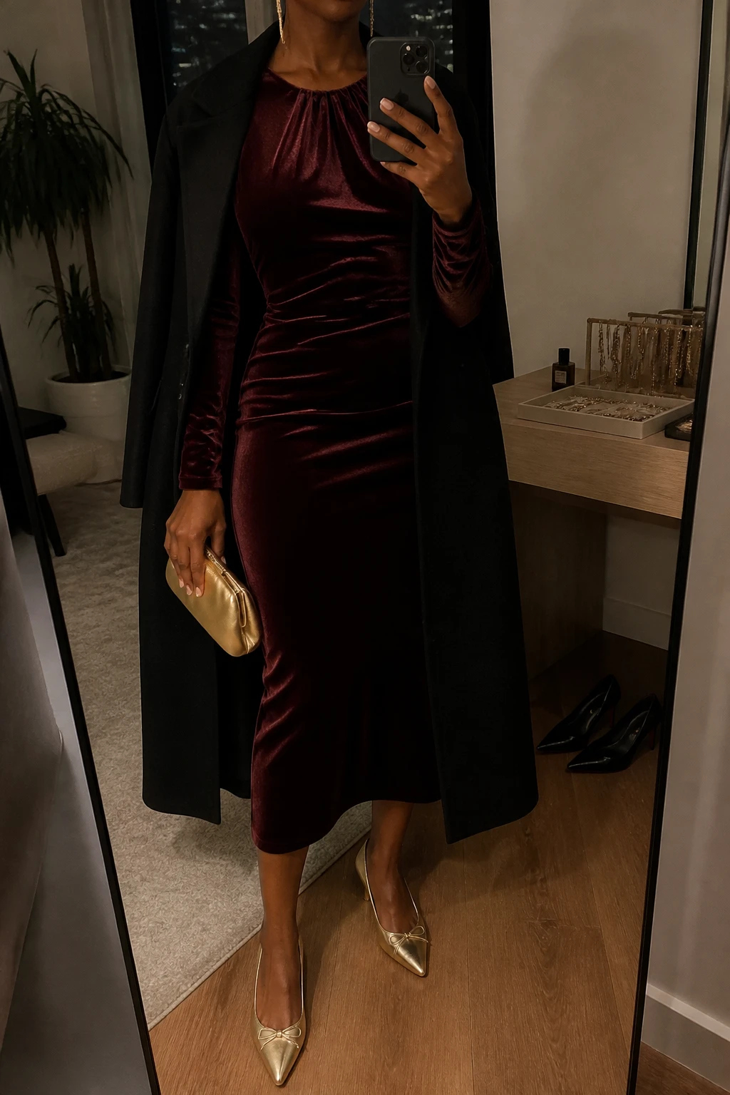 Image Prompt: Mirror selfie of a black woman wearing a burgundy long-sleeve velvet midi dress with a fitted waist and soft gathered neckline, gold metallic pointed-toe kitten heels, a small champagne gold satin clutch held in one hand, delicate gold drop earrings, and a long black tailored wool coat worn open, holding a phone in front of a full-length mirror, cropped at the NECK so the face does not show, with a relaxed casual selfie pose, in a stylish city apartment, warm moody indoor lighting, a jewelry tray on the vanity and a pair of heels on the floor nearby, realistic velvet texture, elegant but natural party styling, latest iPhone photo quality, no additional clothing or accessories.