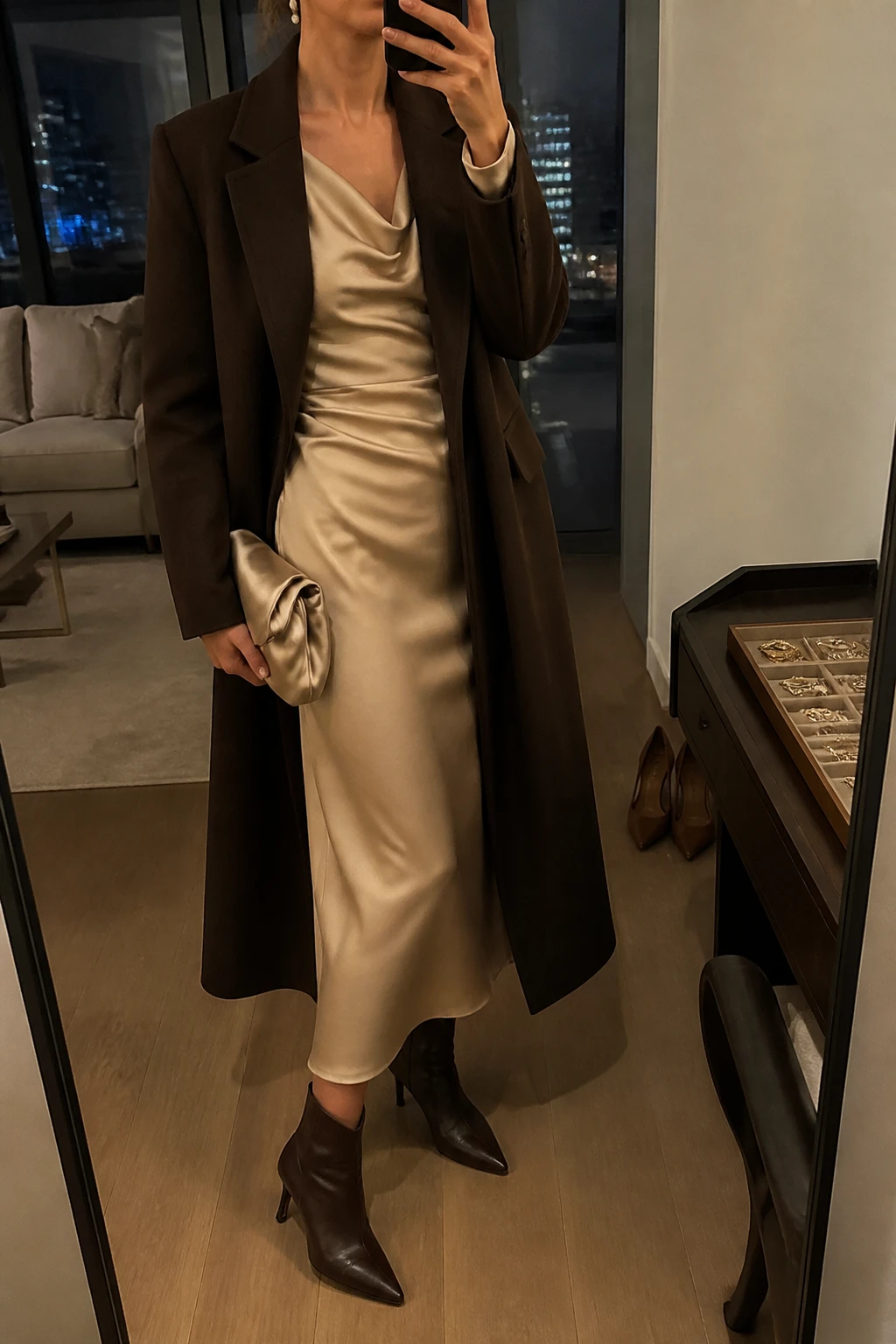 Image Prompt: Mirror selfie of a woman wearing a champagne long-sleeve satin midi dress with a softly draped cowl neckline and gently fitted waist, a long chocolate brown tailored wool coat worn open, dark brown leather pointed-toe heeled ankle boots, a small champagne satin clutch, and delicate gold pearl-drop earrings, holding a phone in front of a mirror, cropped at the NECK so the face does not show, with a relaxed casual selfie pose, in a stylish city apartment, warm moody indoor lighting, a jewelry tray on the vanity and a pair of heels on the floor nearby, realistic champagne satin folds, rich chocolate wool texture and natural evening light, latest iPhone photo quality, no additional clothing or accessories.