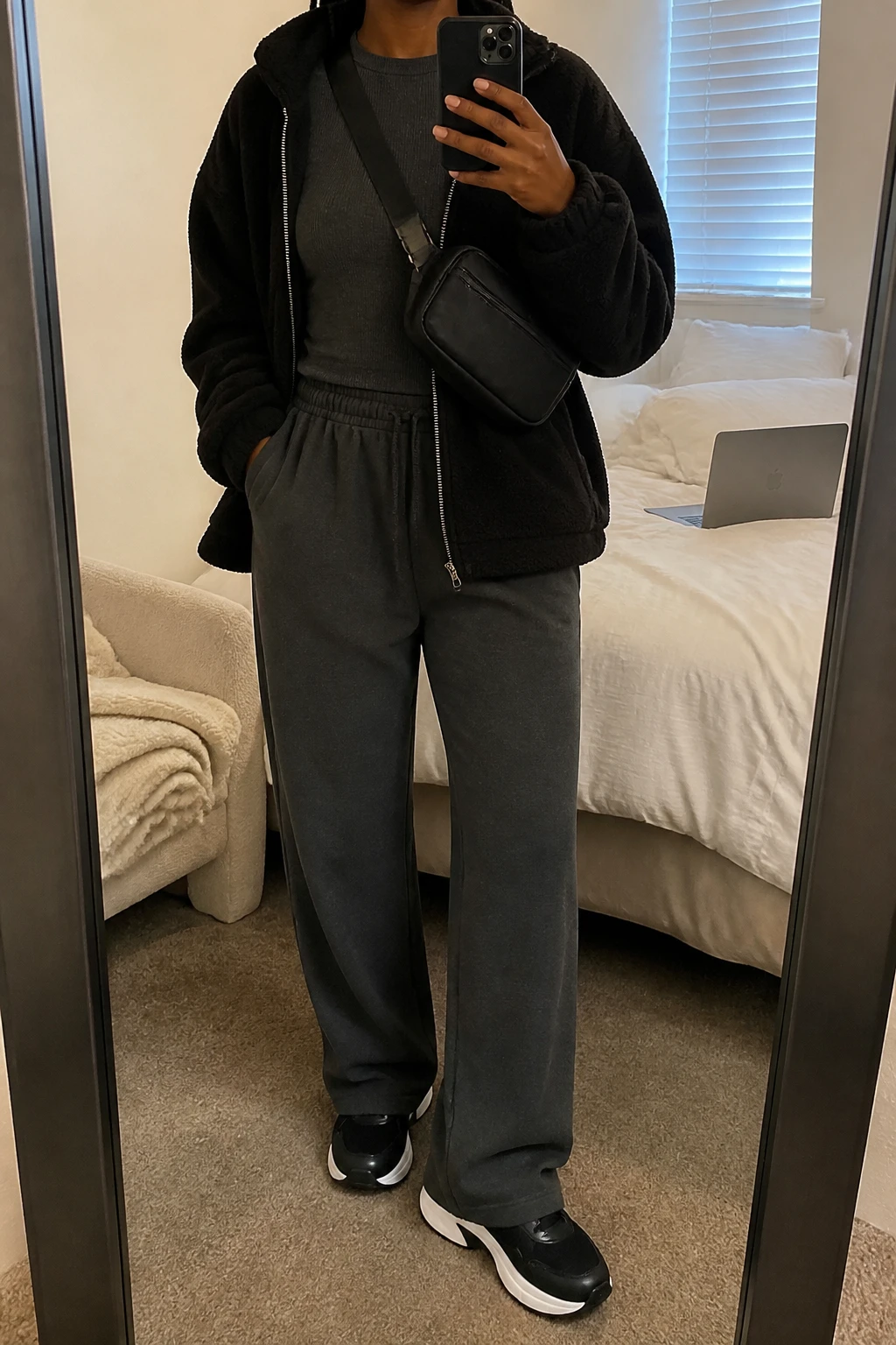 Image Prompt: Mirror selfie of a black woman wearing a black oversized zip-front fleece jacket with a high collar over a charcoal grey fitted thermal crewneck top, matching charcoal grey relaxed straight-leg fleece joggers, black cushioned chunky sneakers with white soles, and a small black nylon zippered crossbody bag, holding a phone in front of a mirror, cropped at the NECK so the face does not show, with a relaxed casual selfie pose, in a cozy bedroom mirror setup, soft indoor lighting, a folded blanket on a chair and a laptop on the bed, realistic black fleece texture, natural charcoal fabric folds and relaxed travel outfit appearance, latest iPhone photo quality, no additional clothing or accessories.