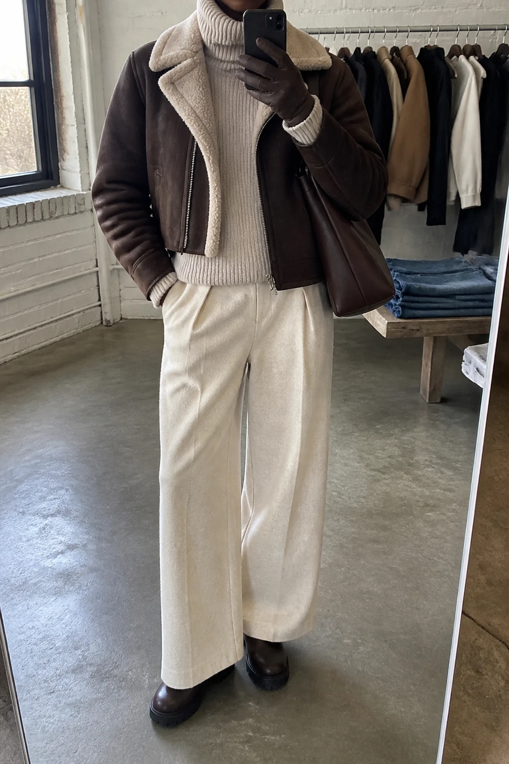 Image Prompt: Mirror selfie of a black woman wearing a dark brown faux-suede shearling-lined jacket with a cream fleece collar over a cream thick ribbed turtleneck sweater, cream wide-leg wool-blend trousers, dark brown waterproof leather chunky ankle boots, a chocolate brown leather shoulder bag, and brown leather gloves, holding a phone in front of a mirror, cropped at the NECK so the face does not show, with a relaxed casual selfie pose, in a fashion studio mirror corner, natural window light, a clothing rack with garments behind and folded denim jeans on a bench, realistic suede and fleece textures, slightly imperfect casual appearance, latest iPhone photo quality, no additional clothing or accessories.