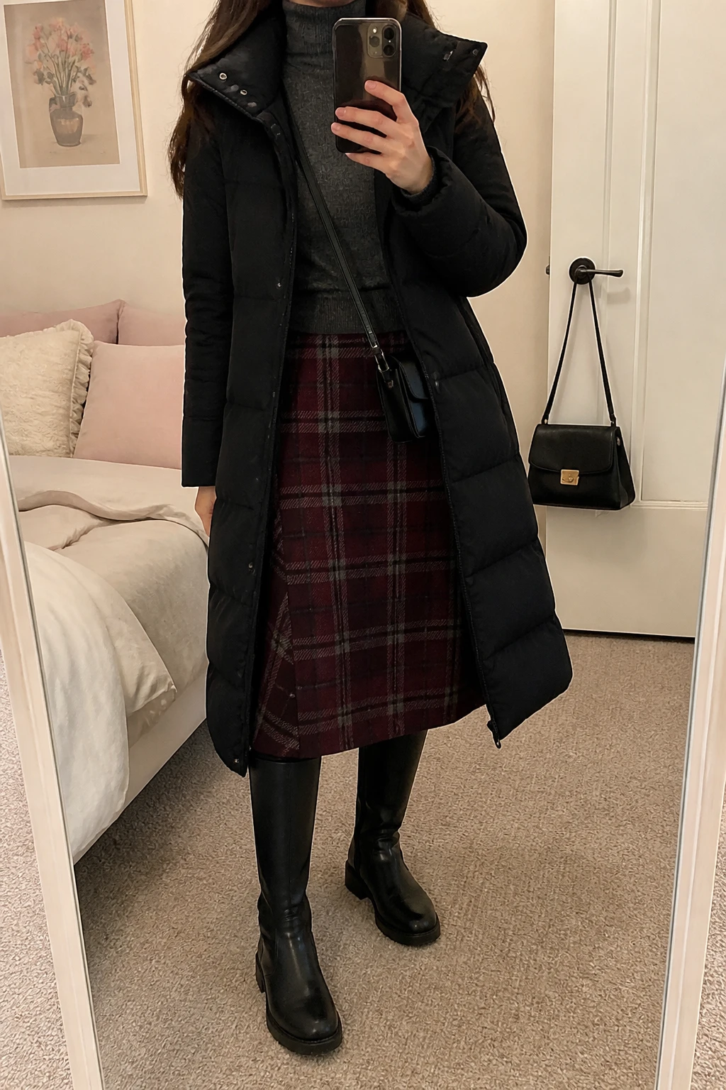 Image Prompt: Mirror selfie of a woman wearing a long black insulated quilted winter coat over a dark grey fitted wool turtleneck sweater, a burgundy and charcoal plaid wool midi skirt, black fleece-lined tights, black leather knee-high flat boots with rubber soles, and a small black structured crossbody bag, holding a phone in front of a mirror, cropped at the NECK so the face does not show, with a relaxed casual selfie pose, in a feminine-themed bedroom, soft indoor lighting, soft pillows on an unmade bed and a handbag hanging on a door handle, realistic plaid texture and natural coat drape, latest iPhone photo quality, no additional clothing or accessories.