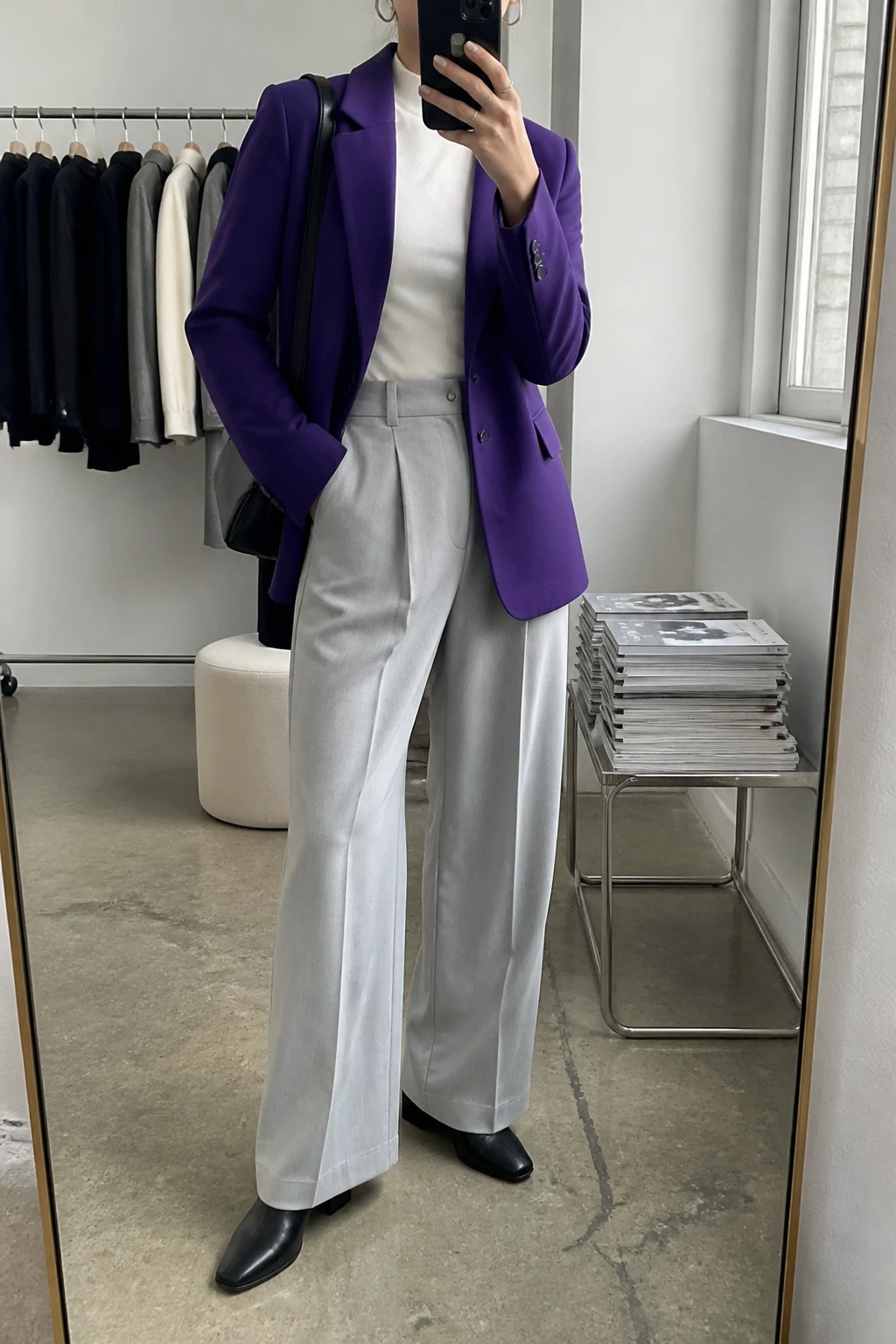 Image Prompt: Mirror selfie of a woman wearing a rich royal purple tailored wool-blend single-breasted blazer over an icy white fitted fine-knit mock-neck sweater, light cool grey high-waisted wide-leg tailored trousers, black leather pointed-toe low block-heel ankle boots, a structured black leather shoulder bag, and small silver hoop earrings, holding a phone in front of a mirror, cropped at the NECK so the face does not show, with a relaxed casual selfie pose, in a fashion studio mirror corner, soft natural window light, a clothing rack with garments behind and a stack of fashion magazines on a side table, realistic royal purple blazer fabric, natural icy grey trouser folds and crisp cool-toned styling, latest iPhone photo quality, no additional clothing or accessories.