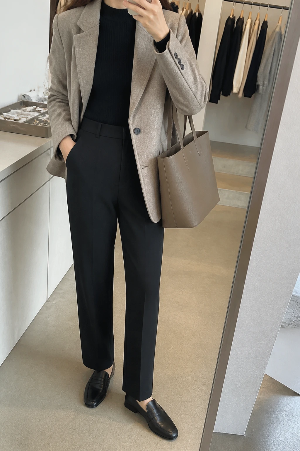 Image Prompt: Mirror selfie of a woman wearing a beige tailored wool-blend blazer over a black fitted ribbed mock-neck sweater, black high-waisted straight-leg tailored trousers, black leather low-heeled loafers, and a structured taupe leather tote bag, holding a phone in front of a mirror, cropped at the NECK so the face does not show, with a relaxed casual selfie pose, in a fashion studio mirror corner, soft indoor lighting, a clothing rack with garments behind and a jewelry tray on the vanity, realistic wool blazer fabric, natural tailored trouser creases, latest iPhone photo quality, no additional clothing or accessories.