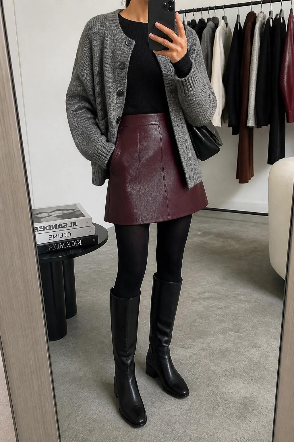 Image Prompt: Mirror selfie of a woman wearing a heather grey chunky button-front knit cardigan over a black fitted long-sleeve thermal top, a burgundy high-waisted faux-leather A-line mini skirt, black opaque fleece-lined tights, black leather knee-high boots with low block heels, and a small black leather shoulder bag, holding a phone in front of a mirror, cropped at the NECK so the face does not show, with a relaxed casual selfie pose, in a fashion studio mirror corner, soft indoor lighting, a clothing rack with garments behind and a stack of fashion magazines on a side table, realistic grey knit cardigan texture, natural burgundy faux-leather skirt finish and warm winter styling, latest iPhone photo quality, no additional clothing or accessories.