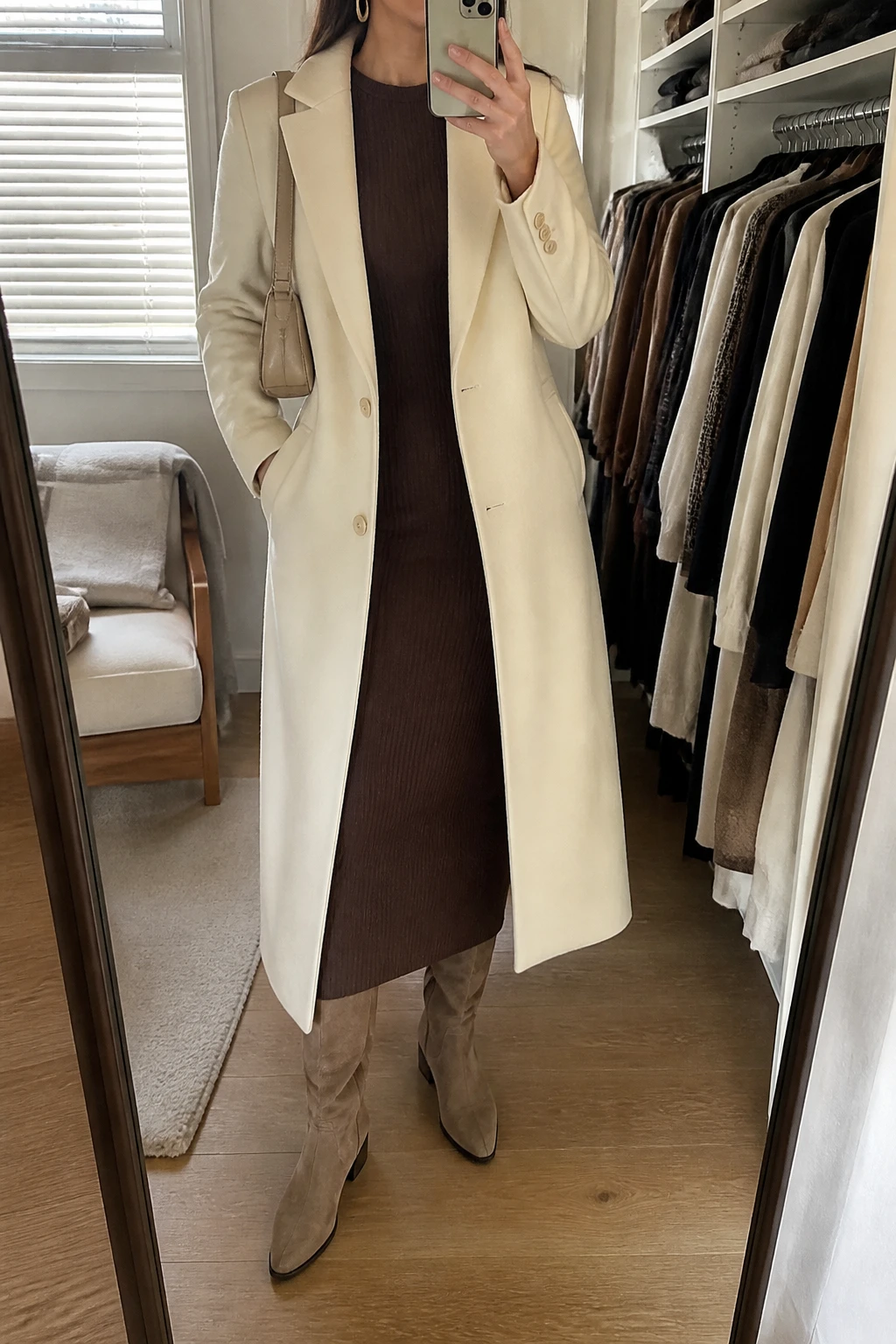 Image Prompt: Mirror selfie of a woman wearing a long cream tailored wool coat over a chocolate brown fitted ribbed knit midi dress, taupe suede knee-high boots with low block heels, a small beige leather shoulder bag, and delicate gold oval earrings, holding a phone in front of a mirror, cropped at the NECK so the face does not show, with a relaxed casual selfie pose, in a walk-in closet mirror, soft natural window light, an open closet with clothes visible and a folded blanket on a chair, realistic taupe suede boots, natural cream wool coat texture and chocolate knit folds, latest iPhone photo quality, no additional clothing or accessories.