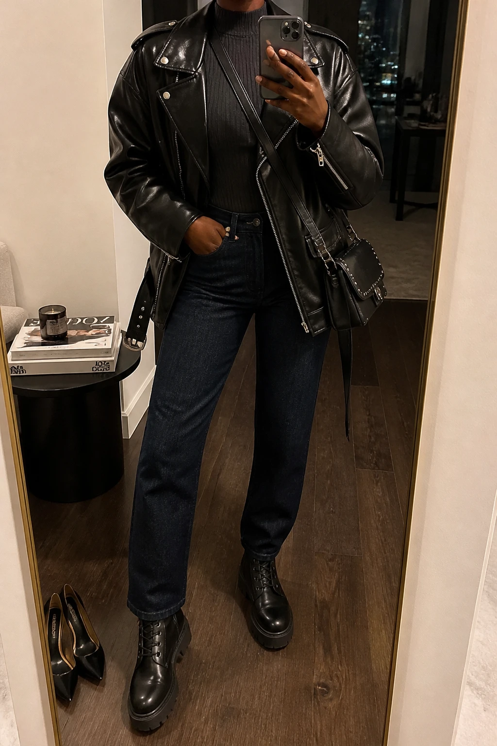 Image Prompt: Mirror selfie of a black woman wearing an oversized black faux-leather biker jacket over a charcoal grey fitted ribbed mock-neck top, dark indigo straight-leg denim jeans, black leather chunky lace-up combat boots with flat rubber soles, and a small black studded leather crossbody bag, holding a phone in front of a full-length mirror, cropped at the NECK so the face does not show, with a relaxed casual selfie pose, in a stylish city apartment, warm moody indoor lighting, a pair of heels on the floor nearby and a stack of fashion magazines on a side table, realistic leather texture, natural denim creases, casual concert-ready appearance, latest iPhone photo quality, no additional clothing or accessories.