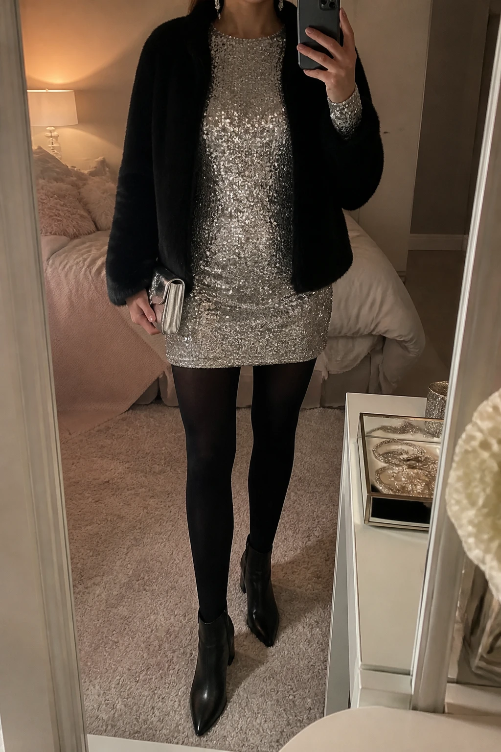 Image Prompt: Mirror selfie of a woman wearing a silver long-sleeve sequin mini dress with a modest round neckline and gently fitted silhouette, black opaque thermal tights, black leather pointed-toe ankle boots with block heels, a cropped black faux-fur coat worn open, a small silver metallic clutch, and delicate silver crystal drop earrings, holding a phone in front of a mirror, cropped at the NECK so the face does not show, with a relaxed casual selfie pose, in a feminine-themed bedroom, warm moody lighting, soft pillows on an unmade bed and a jewelry tray on the vanity, realistic silver sequins with natural subtle sparkle, soft black faux-fur texture and believable evening lighting, latest iPhone photo quality, no additional clothing or accessories.