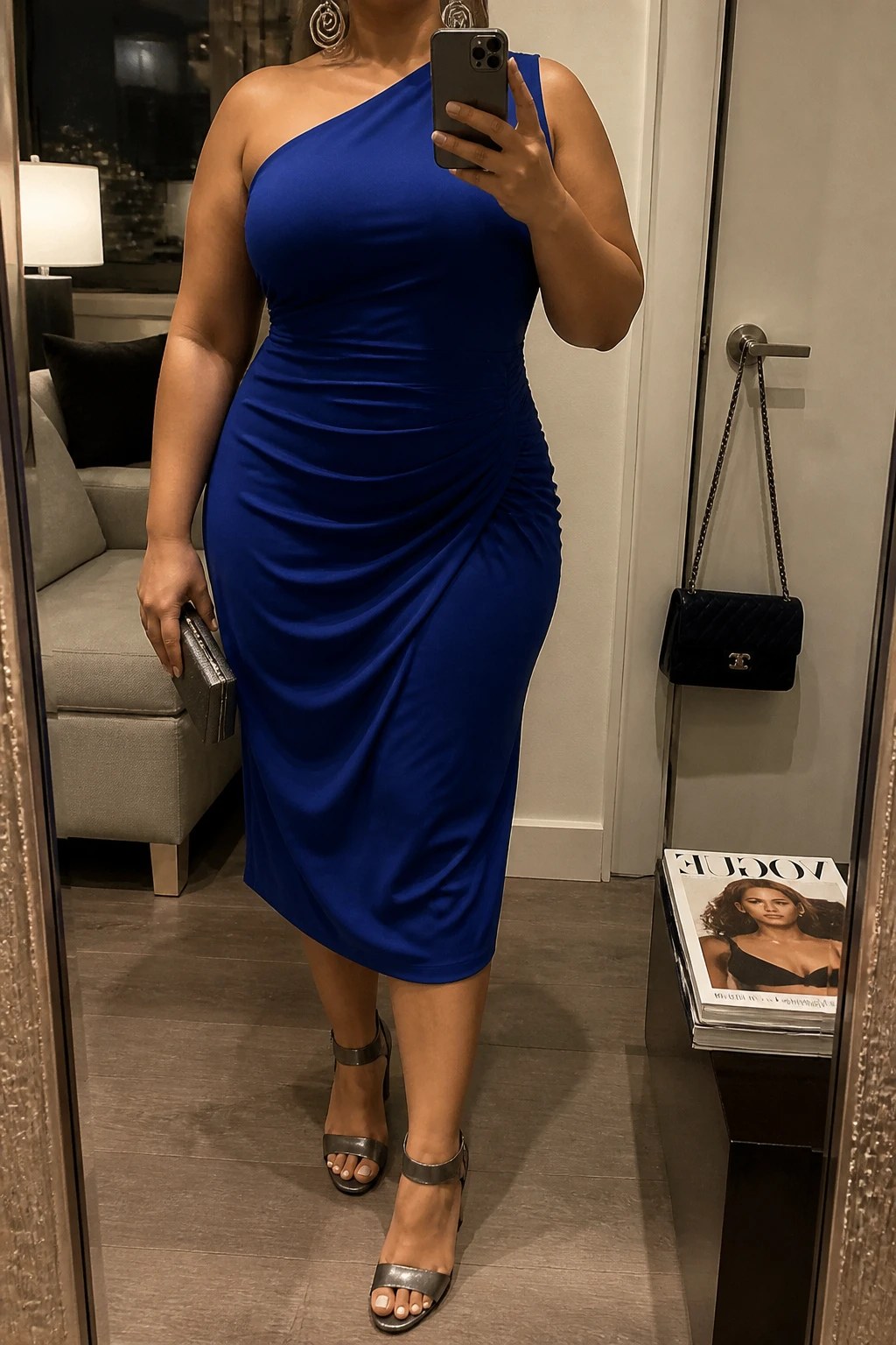 Mirror selfie of a plus size woman wearing a sapphire blue one-shoulder midi dress with gentle waist ruching and a softly draped skirt, metallic gunmetal block-heel sandals, a small gunmetal box clutch, and silver spiral earrings, clearly showing the exact dress shape, sapphire color, ruching, footwear, and accessories listed, holding a phone in front of a mirror, cropped at the neck so the face does not show, relaxed dramatic selfie stance, in a stylish city apartment with warm moody lighting, a handbag hanging on a door handle and a stack of fashion magazines on a side table, realistic graduation party appearance, latest iPhone photo quality.