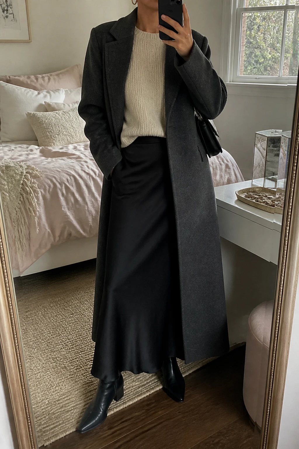 Image Prompt: Mirror selfie of a woman wearing an oatmeal relaxed-fit cashmere crewneck sweater with a small front tuck into a black satin bias-cut maxi skirt, black leather pointed-toe ankle boots with low block heels, a long charcoal grey tailored wool coat worn open, a small structured black leather shoulder bag, and delicate silver hoop earrings, holding a phone in front of a mirror, cropped at the NECK so the face does not show, with a relaxed casual selfie pose, in a feminine-themed bedroom, soft natural window light, soft pillows on an unmade bed and a jewelry tray on the vanity, realistic oatmeal cashmere texture, natural black satin reflections without excessive shine, soft charcoal wool folds, latest iPhone photo quality, no additional clothing or accessories.