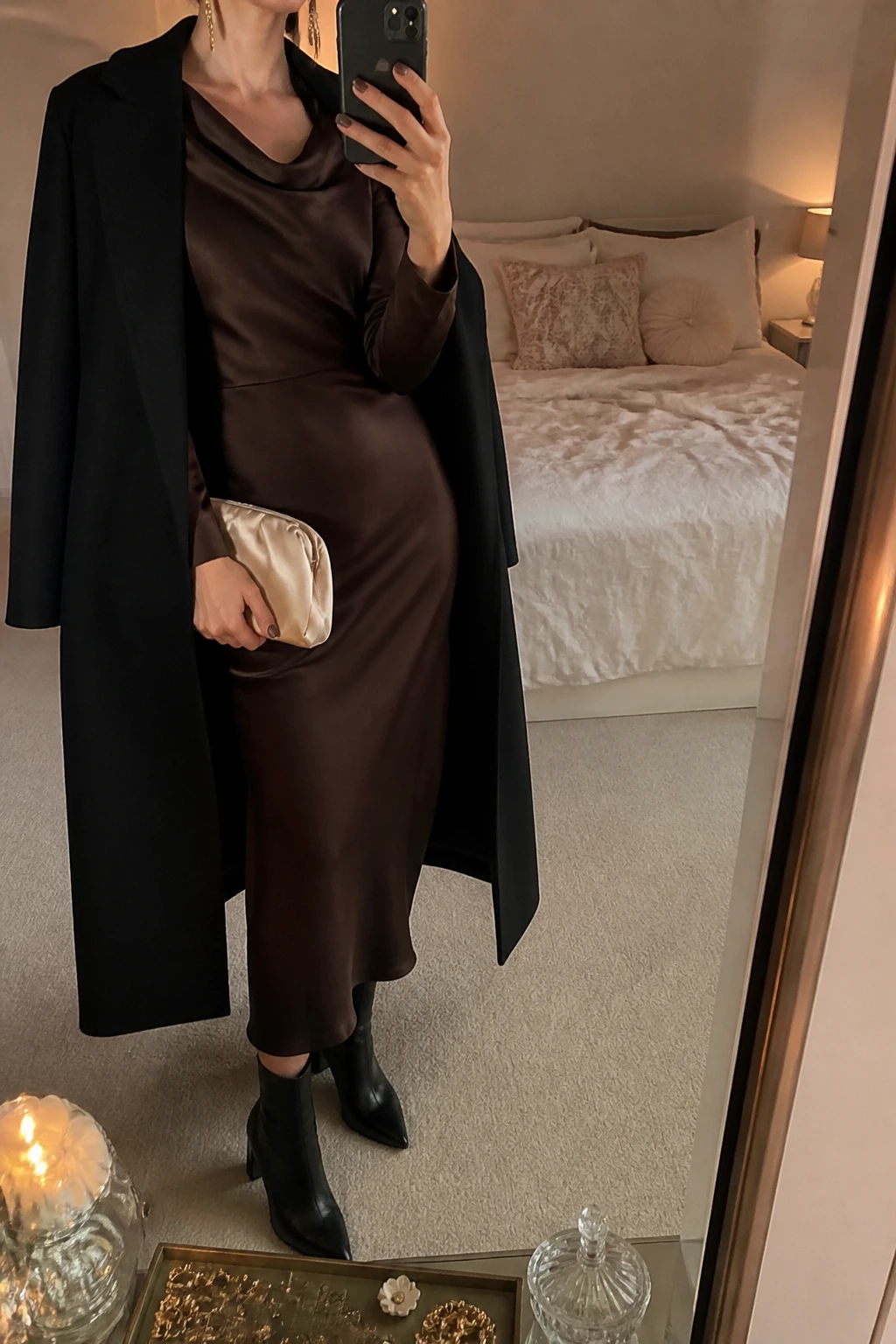 Image Prompt: Mirror selfie of a woman wearing a chocolate brown long-sleeve satin midi dress with a softly draped cowl neckline and gently fitted waist, black leather pointed-toe ankle boots with slim block heels, a long black tailored wool coat worn open, a small champagne satin clutch, and delicate gold drop earrings, holding a phone in front of a mirror, cropped at the NECK so the face does not show, with a relaxed casual selfie pose, in a feminine-themed bedroom, warm moody lighting, soft pillows on an unmade bed and a jewelry tray on the vanity, realistic chocolate satin draping, natural wool coat folds, subtle warm evening light, latest iPhone photo quality, no additional clothing or accessories.