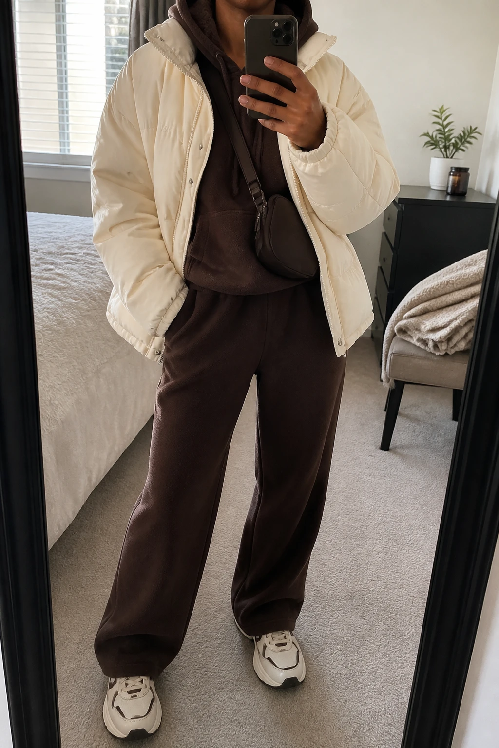 Image Prompt: Mirror selfie of a black woman wearing a cream oversized insulated quilted puffer jacket over a chocolate brown thick fleece-lined hoodie, matching chocolate brown wide-leg fleece sweatpants, cream chunky leather sneakers with brown detailing, and a small dark brown nylon crossbody bag, holding a phone in front of a mirror, cropped at the NECK so the face does not show, with a relaxed casual selfie pose, in a cozy bedroom mirror setup, soft natural window light, a folded blanket on a chair and a small plant on a dresser, realistic cream puffer volume, natural chocolate fleece folds and relaxed winter outfit proportions, latest iPhone photo quality, no additional clothing or accessories.