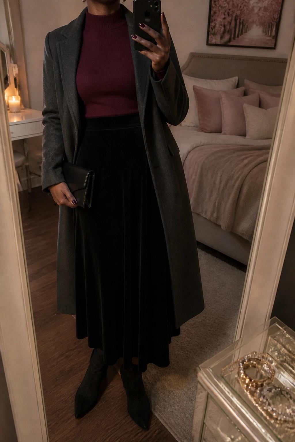 Image Prompt: Mirror selfie of a black woman wearing a deep burgundy fitted cashmere mock-neck sweater tucked into a black high-waisted velvet A-line maxi skirt, black suede pointed-toe ankle boots with low block heels, a long charcoal grey tailored wool coat worn open, a small black satin clutch, and delicate silver crystal stud earrings, holding a phone in front of a mirror, cropped at the NECK so the face does not show, with a relaxed casual selfie pose, in a feminine-themed bedroom, warm moody lighting, soft pillows on an unmade bed and a jewelry tray on the vanity, realistic black velvet texture, natural burgundy cashmere stitches, soft elegant evening shadows, latest iPhone photo quality, no additional clothing or accessories.